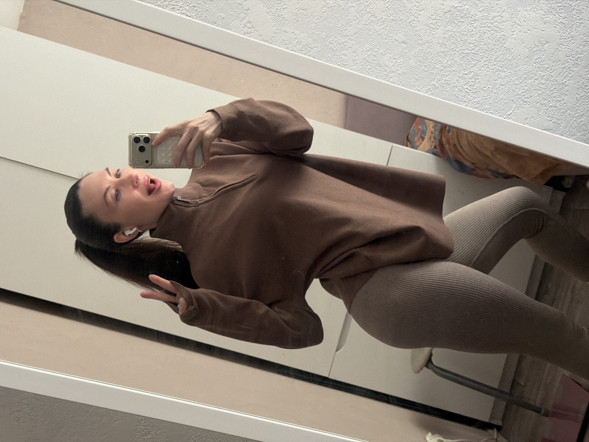 A woman is taking a mirror selfie. She has brown hair pulled back in a ponytail and is wearing a brown sweatshirt and ribbed leggings. She is sticking out her tongue and holding up a peace sign. She appears to be standing indoors, possibly in a room in Perm, Russia, given the presence of what could be furniture and wall texture typical of that region. She is holding a smartphone in her right hand.