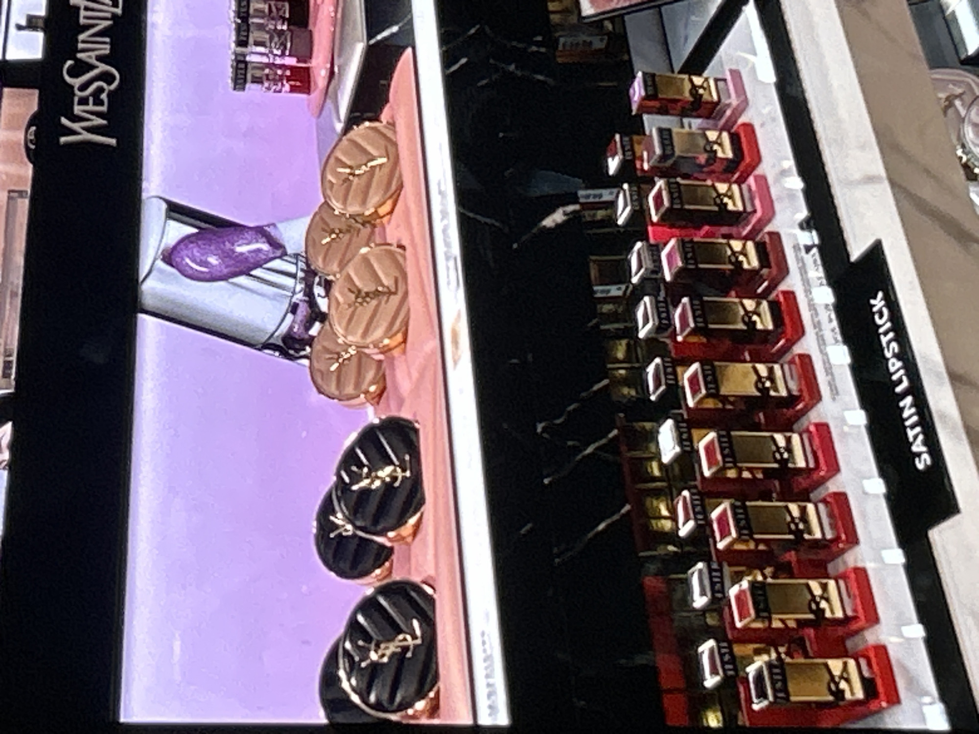 The image displays a retail display of Yves Saint Laurent (YSL) lipstick products, likely in a department store or cosmetics shop. The top of the display features the YSL logo. In the foreground, several rows of YSL lipsticks are arranged on a white surface. These lipsticks are housed in gold-colored tubes with red bases. The word "SATIN LIPSTICK" is visible on the white surface in front of the lipsticks. In the background, there are displays of YSL compacts and lipstick tubes with purple and red contents. The overall environment suggests a well-lit retail setting. The image was taken in Depok, Indonesia, at the Margo City mall.