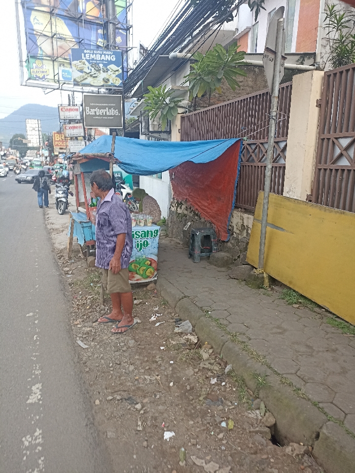 The scene is a street in Jatinangor, Indonesia, with a man standing near a food stall. The man is wearing a blue and white striped shirt and khaki shorts. The stall has a blue and red tarp covering it, and a sign that says "sangs yo" is visible. There are also several items for sale on the stall, including green avocados. The stall is located on the sidewalk, which is made of concrete blocks. There are several power lines running along the side of the building, and the side of the building is painted a yellow color. The man is facing the stall, and he appears to be looking at something on the stall. There are several vehicles parked on the street in the background. There is a green, leafy bush growing on the side of the building. The sidewalk is littered with debris and trash. The street is paved with asphalt. The scene is located at approximately latitude: -6.933 and longitude: 107.773.