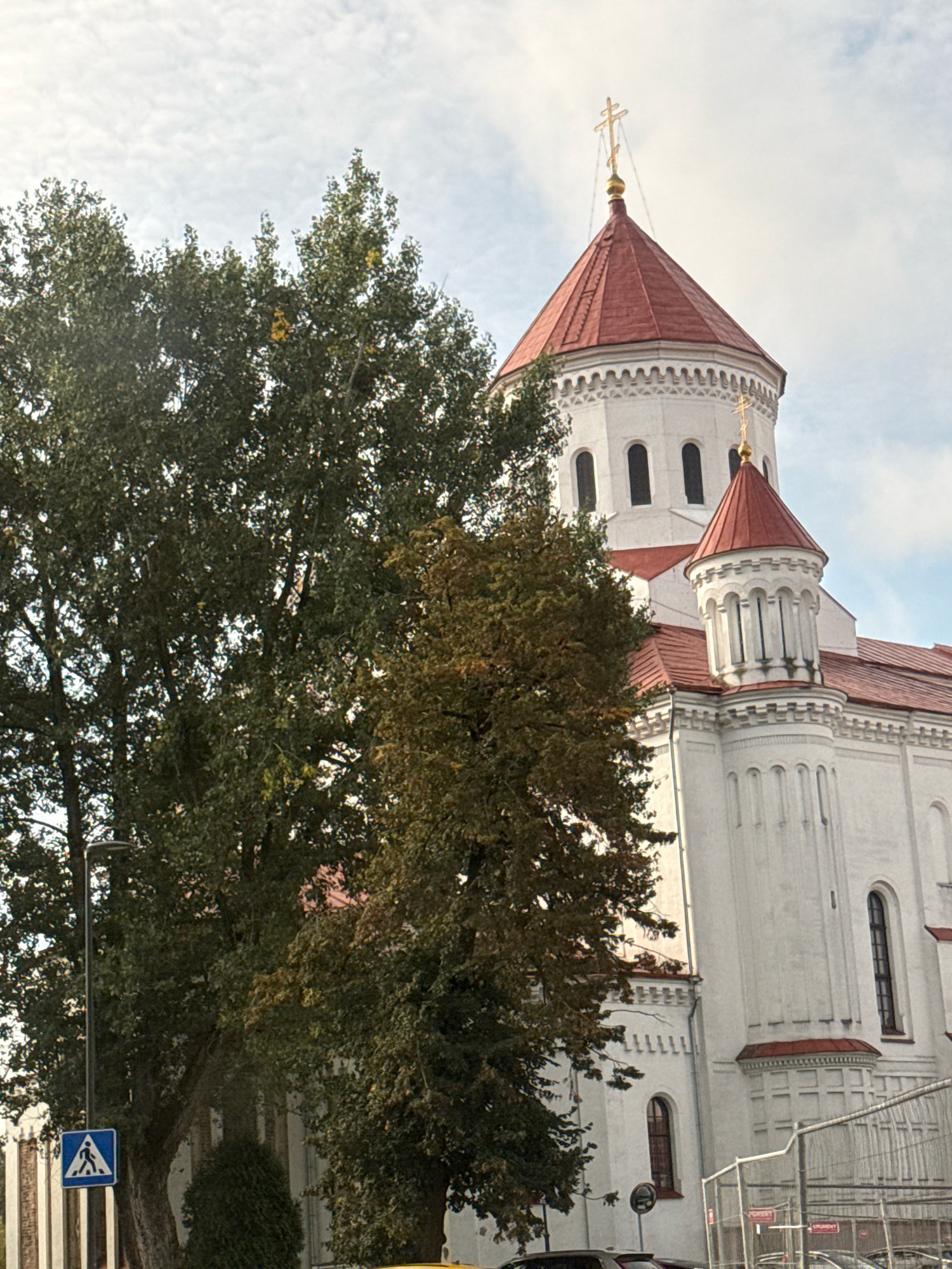 A white Orthodox church with a red roof and gold crosses is partially obscured by trees. A pedestrian crossing sign is visible on the left side of the image, along with a lamppost. A section of a metal fence is present on the right. The sky is cloudy. The image appears to be of the Holy Spirit Orthodox Church in Vilnius, Lithuania.