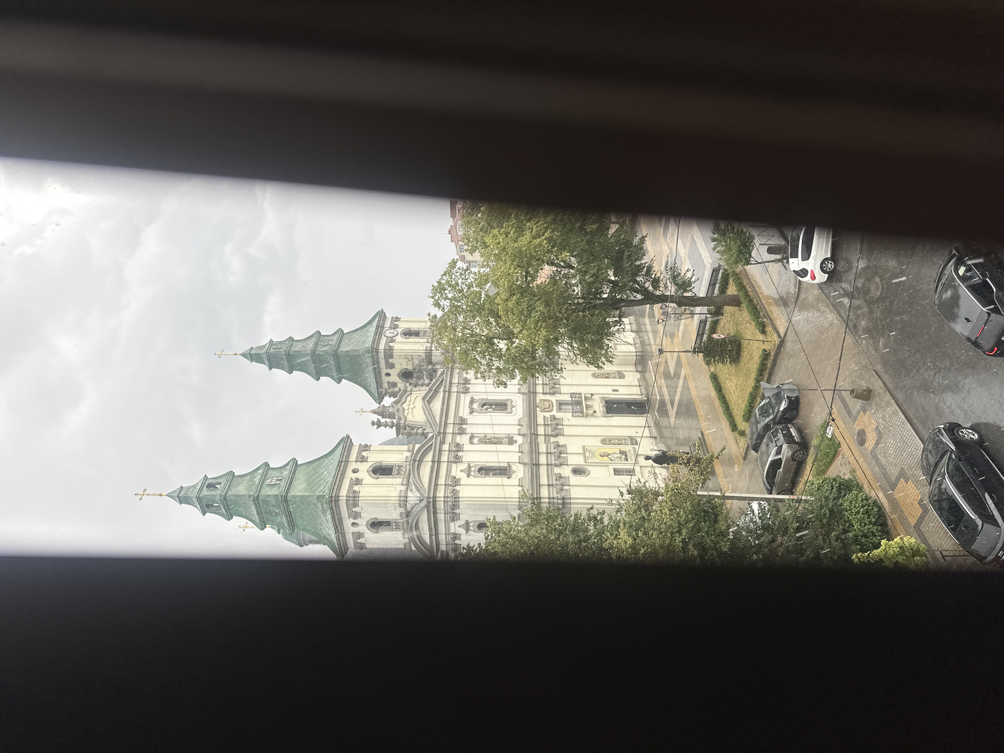 The image depicts a view of the Church of the Immaculate Conception of the Blessed Virgin Mary in Ternopil, Ukraine, seen through a window with dark curtains partially obstructing the view. The church, a prominent landmark, features a striking facade with two green, tiered spires topped with crosses. The sky is overcast, and it appears to be raining, with visible streaks of rain in the air and wet pavement reflecting the light.
In the foreground, several cars are parked on the wet street surrounding the church. Among them are a dark-colored SUV, a silver hatchback, and a black sedan. A white hatchback is visible further down the street. The area around the church is paved with cobblestones and has patches of green grass and trees. A statue of a person in a dark coat stands near the church entrance. The overall scene suggests a cloudy and rainy day in a city square.