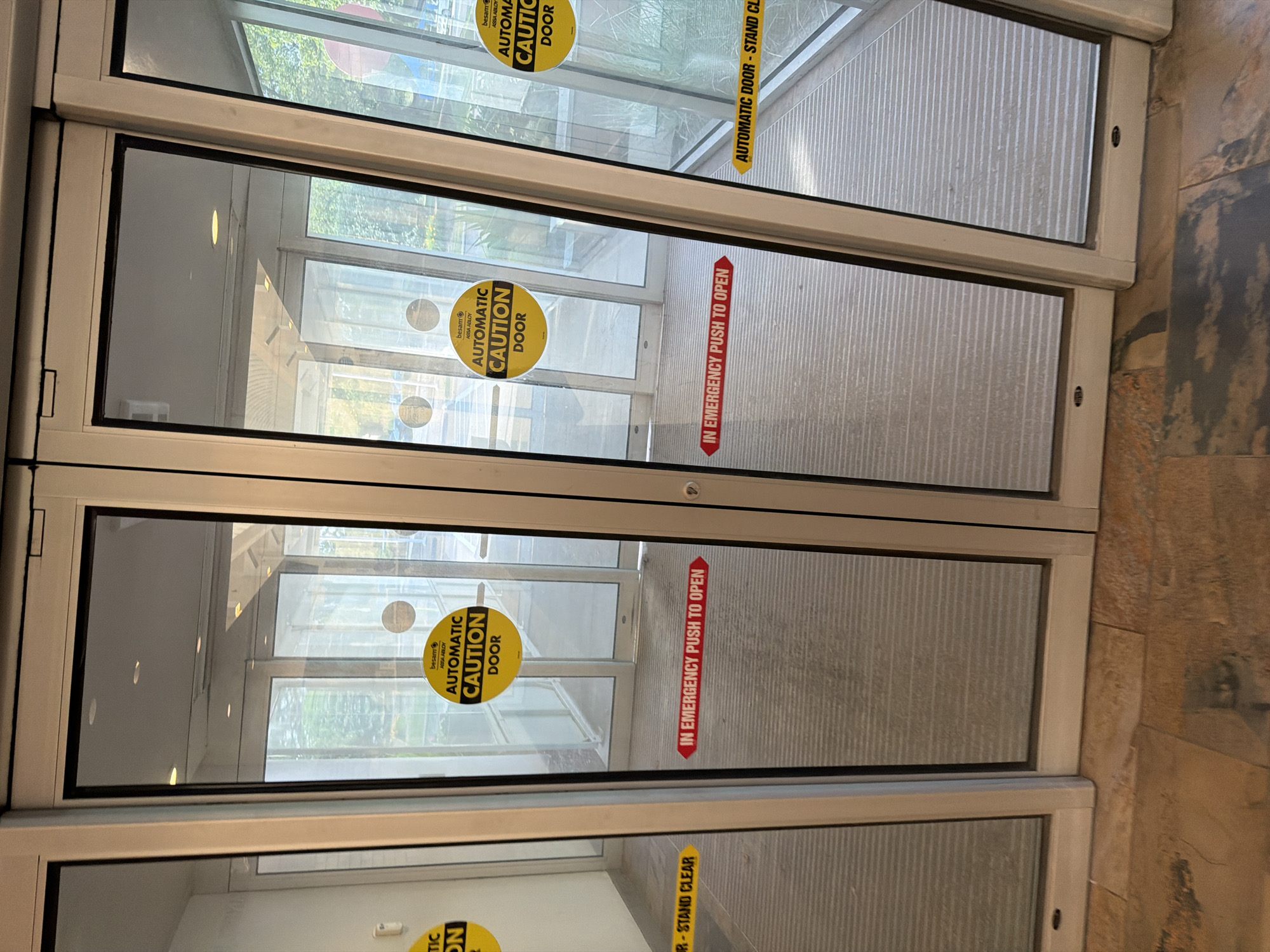 The image shows a set of automatic glass doors at the entrance to a building. The doors are clear glass with black frames and are divided into multiple panels. There are several yellow circular stickers on the glass that read "AUTOMATIC CAUTION DOOR". There are also red rectangular stickers that say "IN EMERGENCY PUSH TO OPEN" and yellow rectangular stickers that say "AUTOMATIC DOOR - STAND CLEAR". The floor in front of the doors is tiled with a pattern of brown and grey stones. The background shows a hallway or interior space with more glass doors and a reflection of trees and sky. The image was taken in Beachwood, United States of America.
