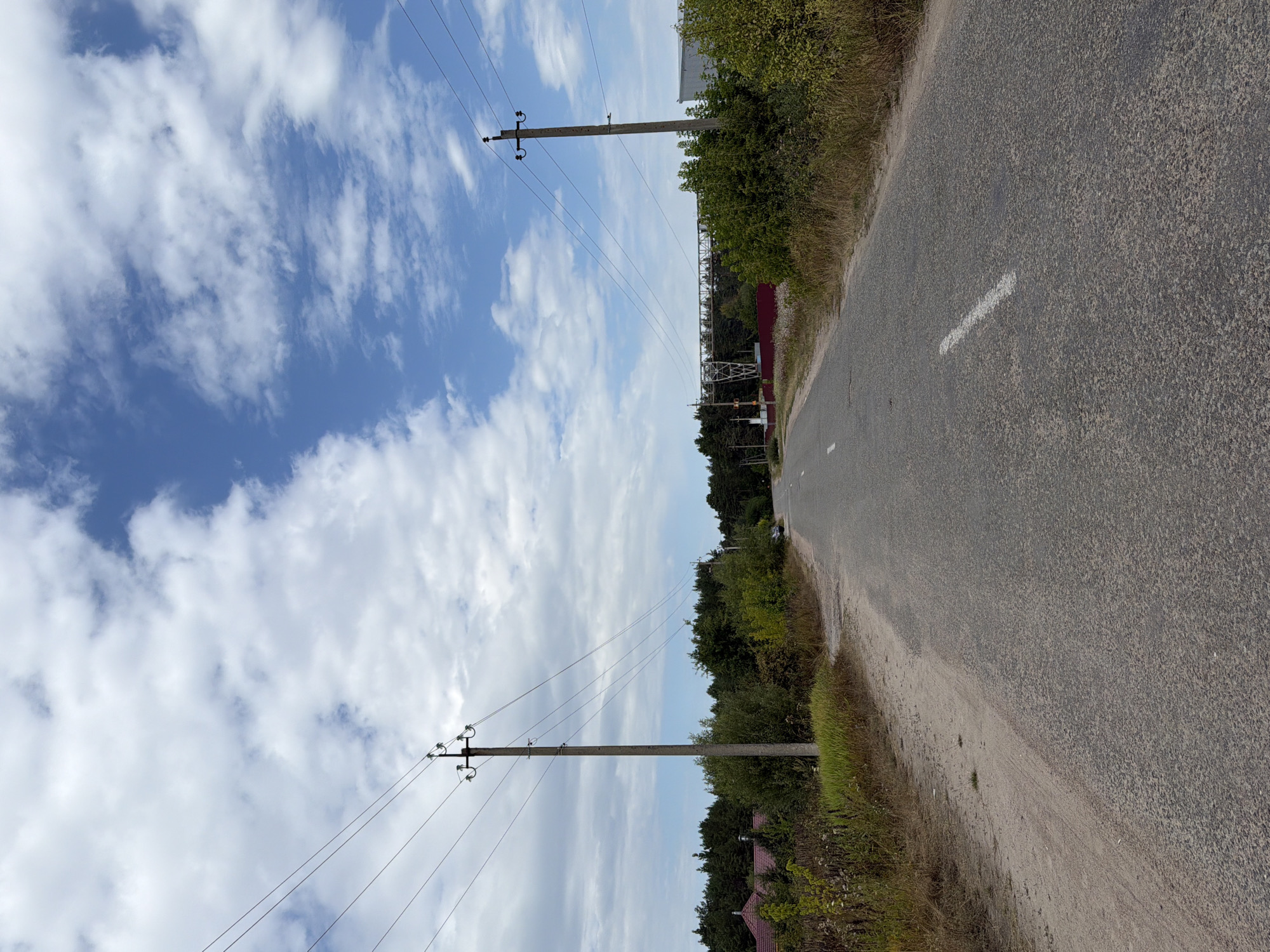 An overcast sky with white and blue clouds looms over a paved road in Mali Kosharyshcha, Ukraine. Two utility poles with power lines straddle the road. The left pole is closer to the foreground, while the right pole is further down the road. The road is in disrepair with patches and cracks. Trees and vegetation line both sides of the road. In the distance, there is a red-roofed building on the left, and a larger industrial-looking structure on the right. A car is parked on the right side of the road in the distance. The scene appears to be rural, possibly on the outskirts of a village.