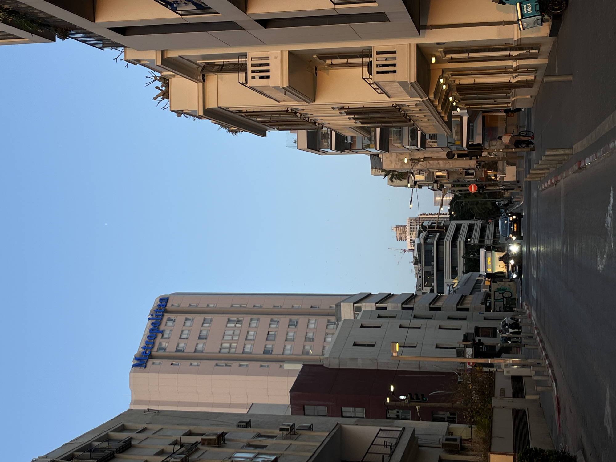 The image depicts a street scene in Tel-Aviv, Israel. The view is looking down a straight street flanked by buildings on both sides. On the left side, there is a tall building with the blue "Metropolitan Hotel" logo on the upper part. There is another building in front of it with air conditioning units visible on the bottom part. On the right side of the street, there are older, light-colored buildings with balconies. The street is paved, and there is a car with its headlights on approaching. There are also several parked motorcycles and bikes on the right side of the road. The sky is clear and blue.