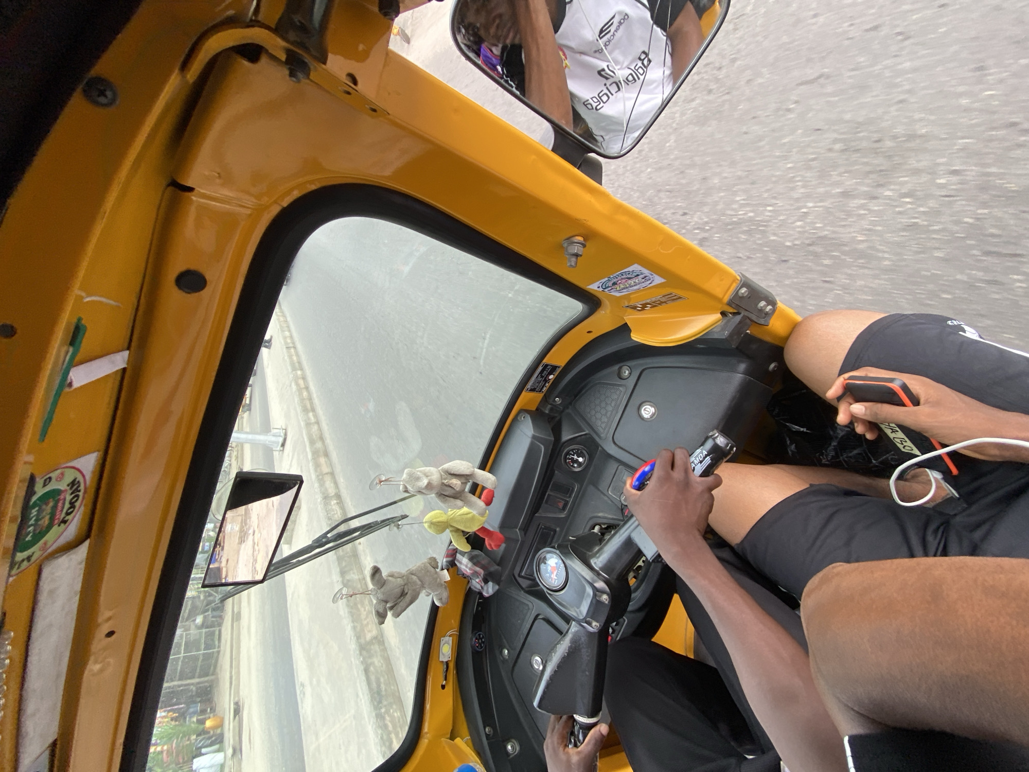 The image, taken from inside a yellow-colored commercial vehicle, appears to be in the city of Ado, Nigeria. The driver, holding a steering wheel, is a person of color, wearing black clothing, and is handling a dark-colored object, possibly a beverage bottle. Beside the driver, there is another person of color wearing black shorts, holding a mobile phone. The windshield is equipped with wiper blades and hanging decorative plush toys. A side mirror shows a reflection of a person wearing a white and black shirt. Outside the vehicle, there's a view of the city with roads and buildings visible. A sticker is visible on the vehicle's interior, bearing the word "Ado".