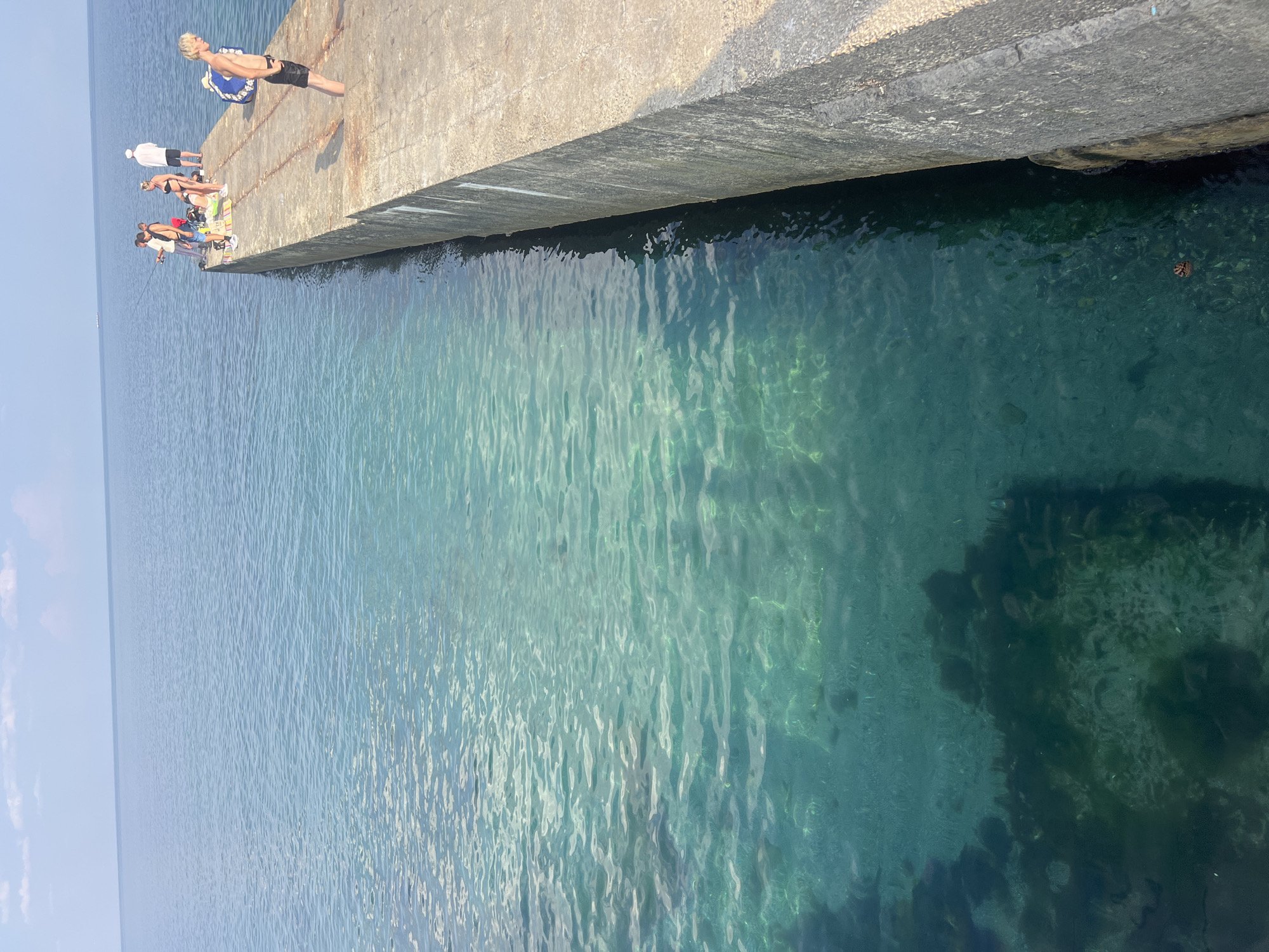 The image depicts a sunny day at the sea with a concrete pier extending into the water. The water is clear and turquoise, with the seabed visible in some areas. Several people are present on the pier. In the foreground, a young man with blond hair is standing, looking out at the sea. Further down the pier, a group of people, including two men fishing with rods, a woman in a bikini, and another person in white, are gathered. A blue float lies on the pier near the young man. The horizon shows a calm sea under a partly cloudy sky, with a distant boat visible. The scene appears to be on a pier in Odesa, Ukraine, a popular coastal city known for its beaches and maritime activities.