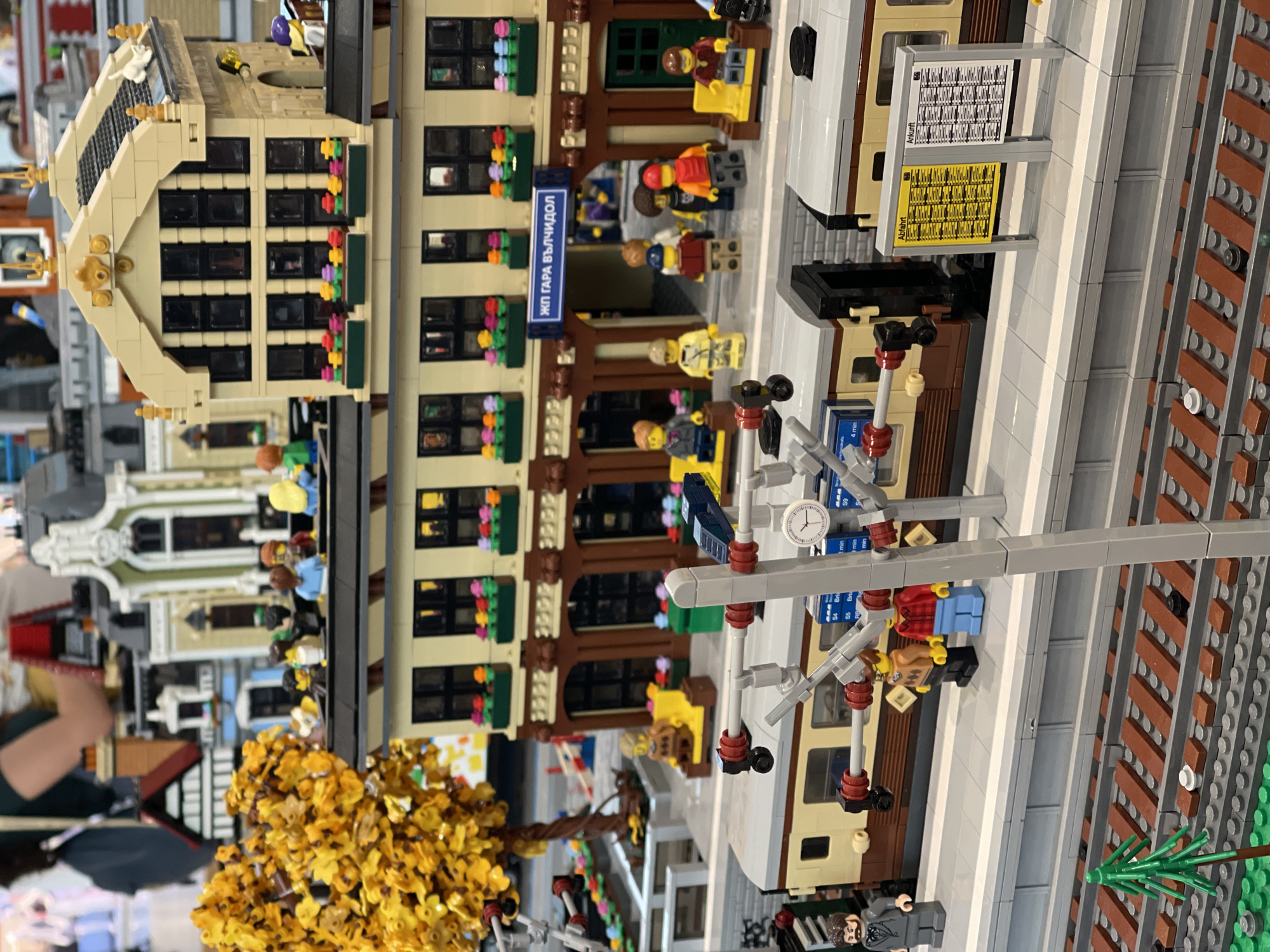 This image depicts a detailed Lego model of a train station and its immediate surroundings, viewed from a slightly elevated perspective.
The central subject is a multi-story Lego train station building constructed predominantly with tan and dark brown bricks. The lower level features several arched windows and a central arched entrance, above which a blue sign displays the text "ЖП ГАРА ВЪЛЧИДОЛ," identifying it as Valchidol Train Station in Bulgaria. The upper levels of the building are tan, with numerous black-framed windows, each adorned with window boxes containing assorted colorful Lego flowers (red, orange, pink, purple, yellow, green). A more elaborate, gabled tan building section with multiple windows sits atop the main station's lower roof. In the background, other blurred Lego buildings are visible, including one with a black and white half-timbered design.
To the left of the station building, a large Lego tree with dense yellow and orange foliage signifies autumn. A light gray Lego platform runs along the front of the station, where two tan and dark brown Lego train cars are parked on a set of brown-tied tracks. In the foreground, two parallel sets of Lego train tracks are visible, with a small green Lego pine tree positioned beside them. Above the platform and tracks, a gray Lego catenary or signal gantry structure is present, featuring red and black connectors and a small white clock face. On the platform, two information boards stand side-by-side: a yellow board labeled "Abfahrt" (Departure) and a white board labeled "Ankunft" (Arrival), both displaying grids of black text.
Numerous Lego minifigures populate the scene, simulating a busy station environment. Minifigures are positioned on the platform, some seated on yellow benches, others standing or walking. A minifigure in a gray suit and black beard is visible on the far left of the platform. Several minifigures are on the lower roof of the main station building. Minifigures are also seen near and within the arched station entrance, including one in a red shirt, one in a blue shirt, and one in a yellow jacket. The minifigures' poses suggest various actions such as waiting, observing, and commuting.