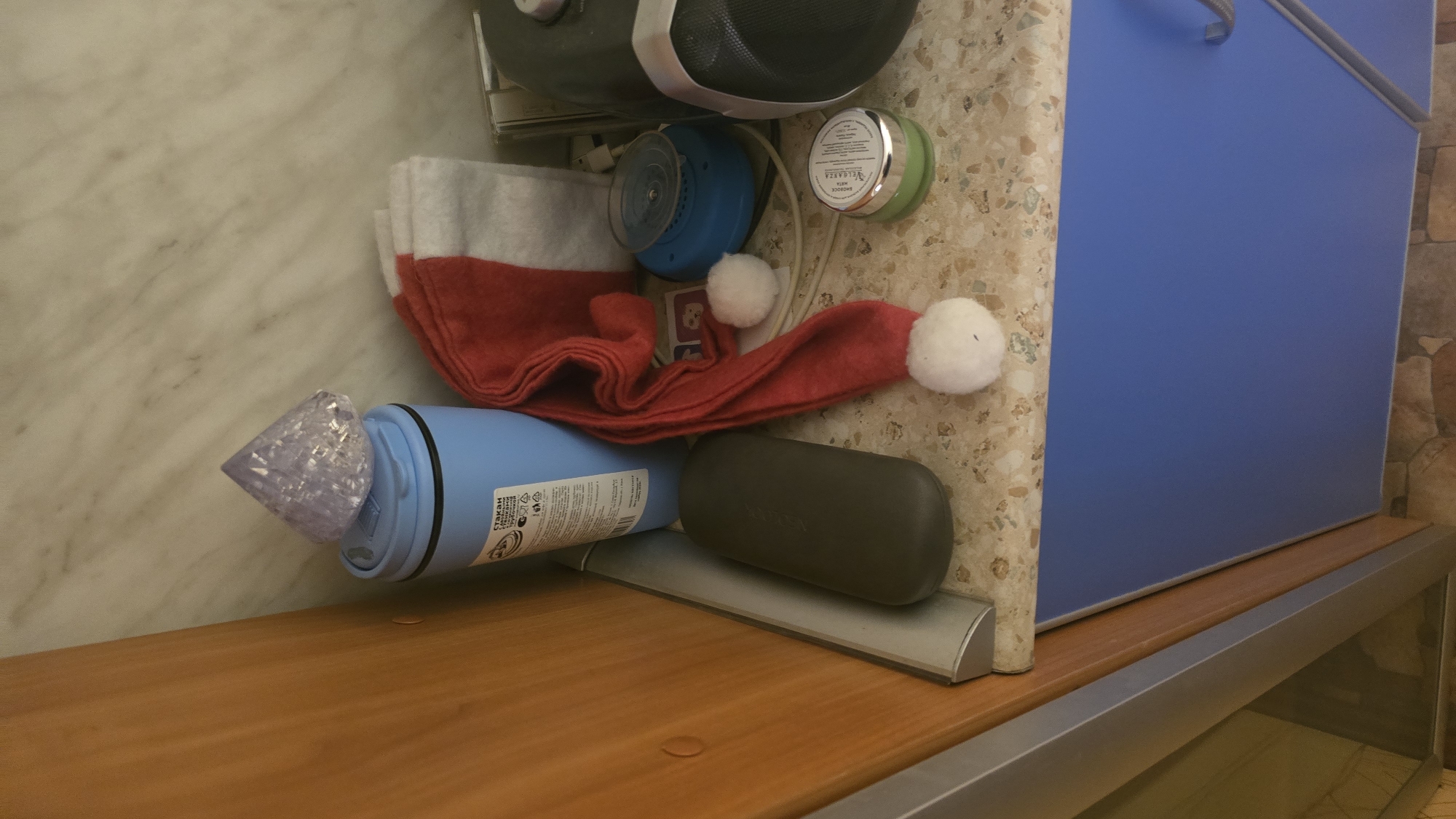 The image depicts a cluttered shelf or countertop. On the left, a wooden cabinet is visible. A light blue cylindrical container with some text on its side sits on the surface. On top of the blue container is a faceted, translucent purple object. To the right of the blue container, a red Christmas hat with white pom-poms is draped, and to its right, a stack of white folded items, possibly towels or cloths. A small blue circular object with a grid pattern is next to the hat, and behind it, wires are visible. Further to the right, a green circular container with a silver lid is present, and a dark grey object, possibly a speaker, is on the far right. The surface appears to be made of a speckled material. A blue cabinet is visible below the shelf. The scene does not provide any information that would allow for the determination of its location in Ukhta, Russia.