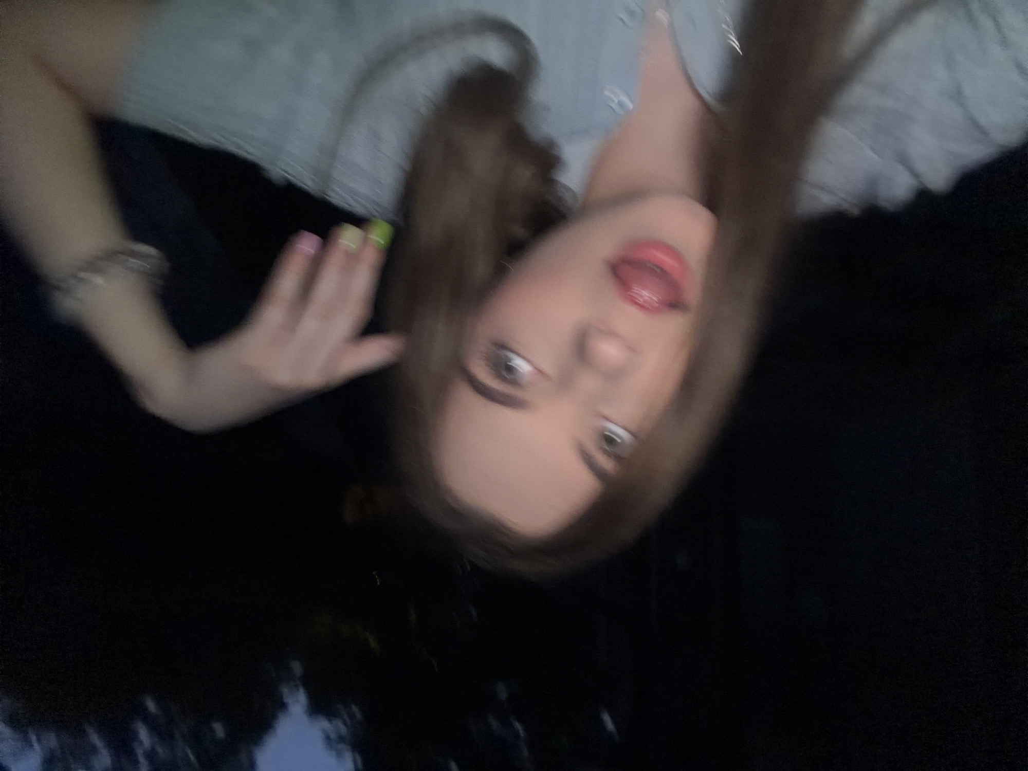 A young woman with long, brown hair is captured in a selfie taken outdoors at dusk or night. She is wearing a light blue collared shirt and has her tongue sticking out, with her mouth slightly open. Her eyes are looking upwards and to the right. Her left hand is raised, with fingers curled, and her fingernails are painted with two different colors, green and pink. A bracelet is visible on her wrist. The background is dark and out of focus, suggesting trees or foliage. There is no specific indication of the city of Dnipro, Ukraine, in the image.