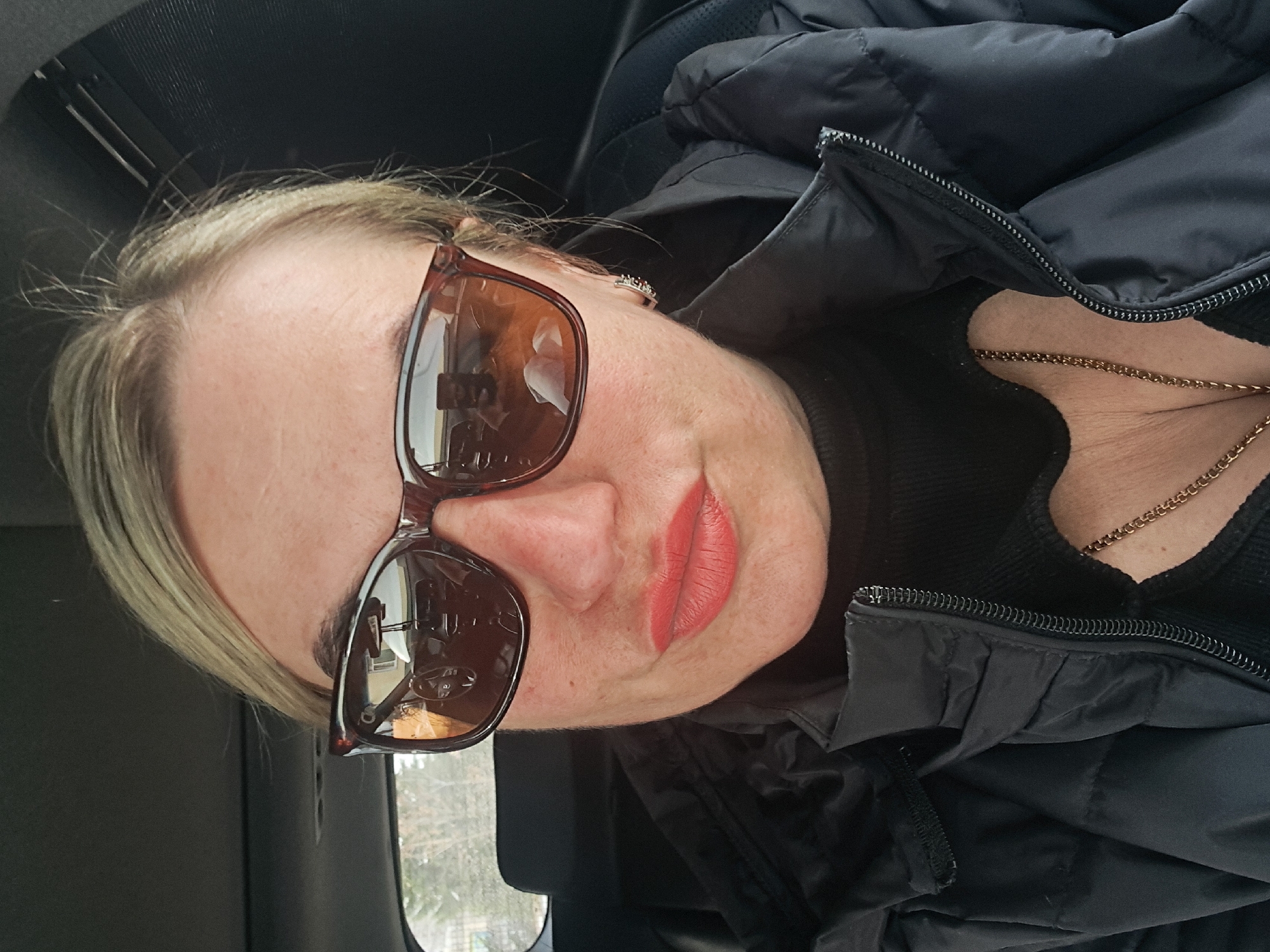 A close-up selfie depicts a woman seated in the passenger compartment of a vehicle in Elektrostal, Russia. The woman has blonde hair, a fair complexion with visible freckles, and wears coral-red lipstick. She is wearing brown-framed sunglasses with reflective lenses, which obscure her eyes. Her attire includes a black quilted puffer jacket, unzipped to reveal a black ribbed top with a heart-shaped cutout at the chest. A gold-colored chain necklace rests against her sternum. A small, silver-toned earring is visible on her right earlobe. Reflections within the sunglasses reveal the vehicle's interior, including a steering wheel, dashboard, and a visible smartphone held by a hand, consistent with taking a selfie. Beyond the vehicle's windshield, a white building with dark windows and sparse tree branches are discernible, indicating an outdoor setting, likely a street or parking area.