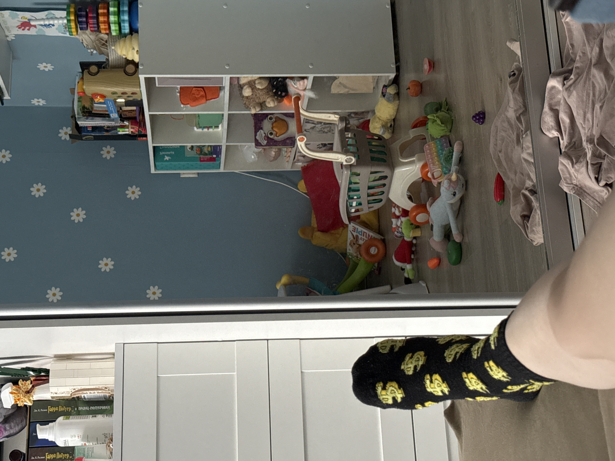 The image shows a cluttered playroom or children's bedroom. On the left, a wall painted light blue with white daisy patterns is visible, and a white shelving unit filled with toys and books stands against the wall. A small trolley filled with more toys sits in front of the shelves.
On the right side of the image, there is a section of what appears to be a mirrored or glass sliding door, and some rumpled tan-colored clothing. In the foreground, a person is partially visible, wearing black socks patterned with yellow dollar signs and a khaki-colored garment.
A portion of Russian text from a book is visible in the image.