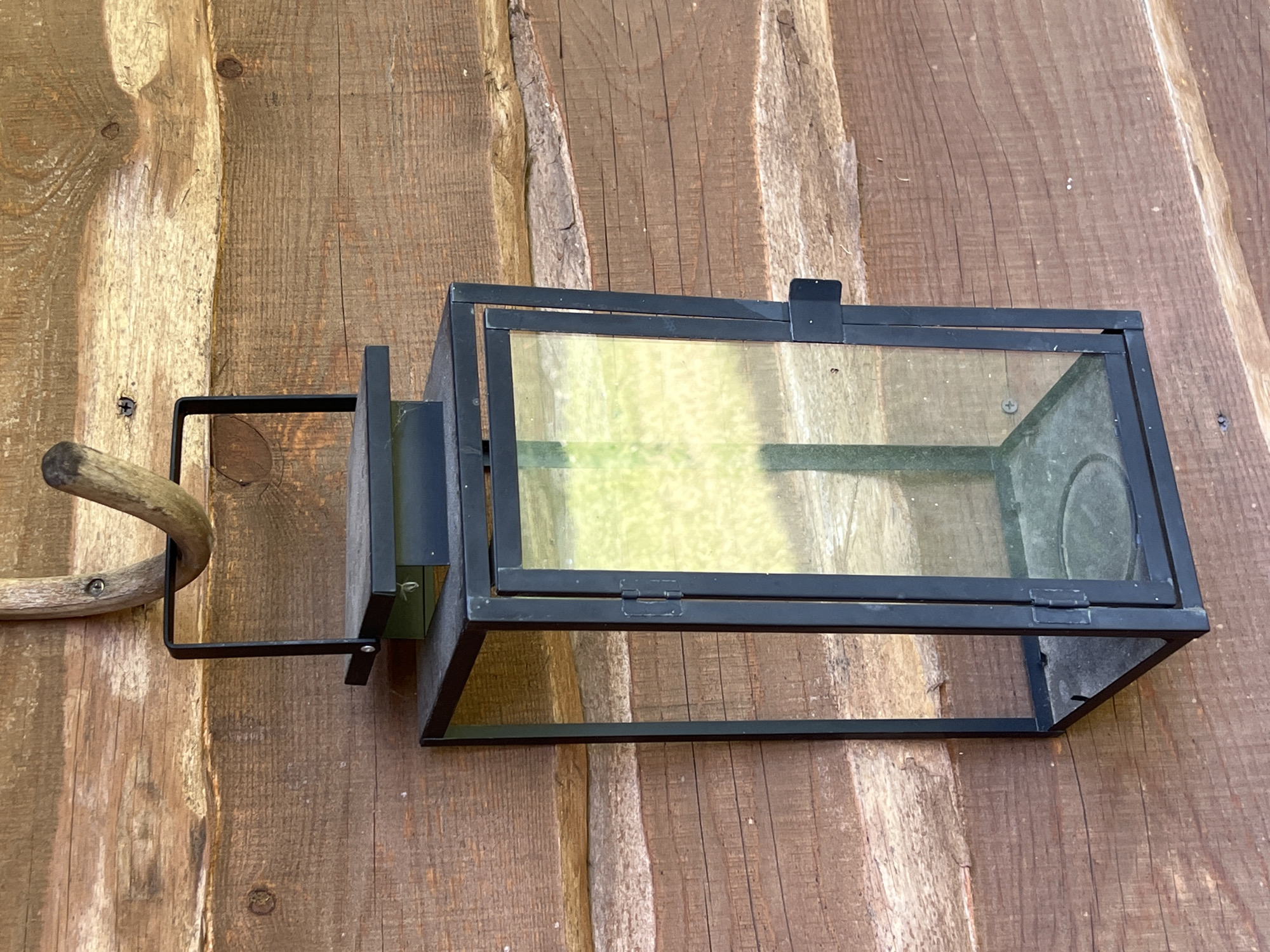 An outdoor scene in Norviliškės, Lithuania features a black metal lantern hanging from a curved, brown wooden beam against a backdrop of weathered wooden logs. The lantern is rectangular with glass panes on its sides, reflecting a blurry, green, and yellow outdoor environment. It has a black handle at the top for hanging and a latch on the right side. The wooden logs are horizontally arranged, showing varying shades of brown and distinct grain patterns, with visible knots and rough textures. The lighting suggests daylight.
