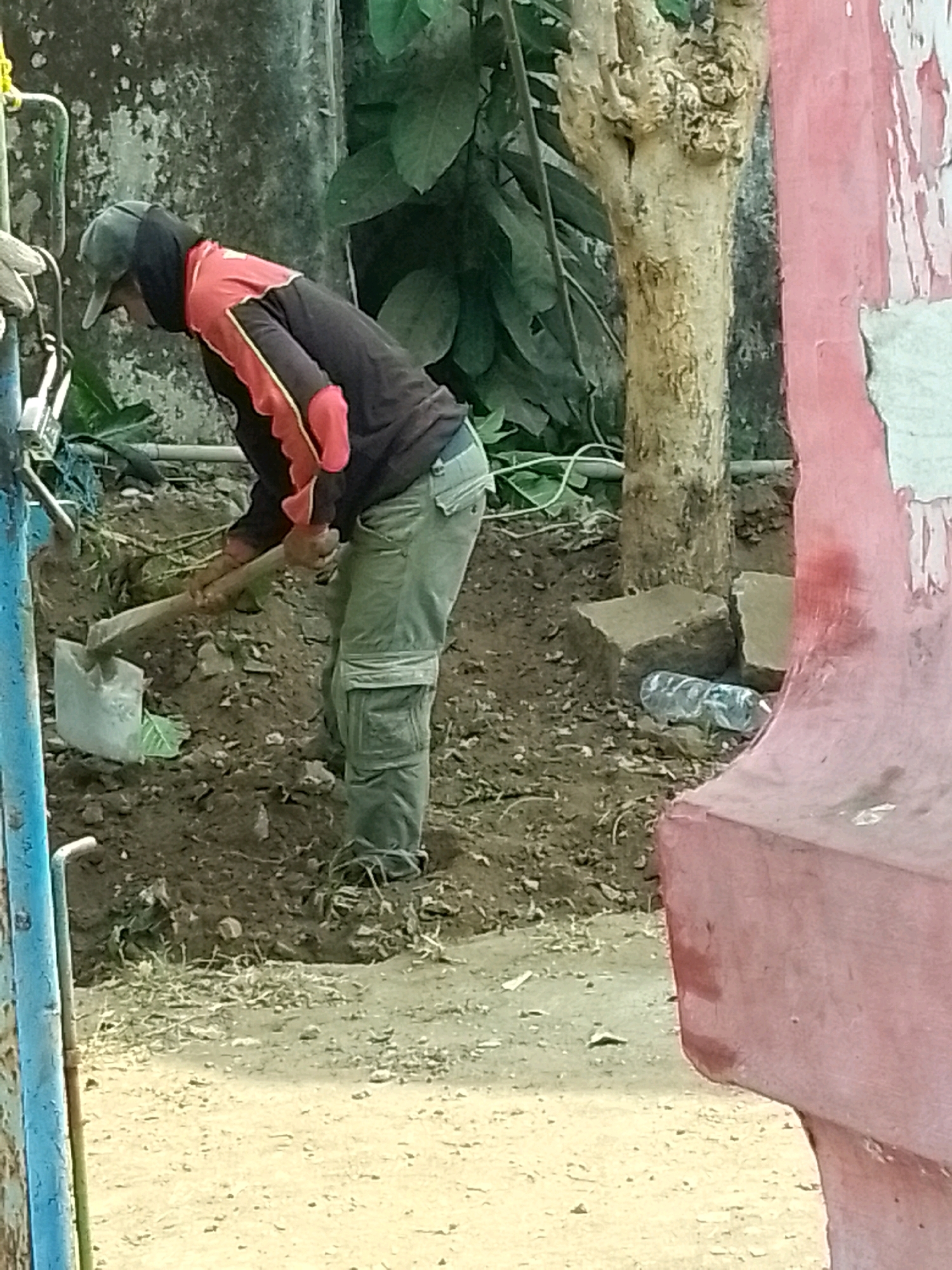 The image depicts a person in work clothes digging in a patch of soil. The person is wearing a dark hooded jacket with red and yellow accents, light green cargo pants, and a dark baseball cap. They are actively using a shovel, which is partially embedded in the dirt. The surrounding environment includes loose soil, some dry grass, a small tree trunk, large concrete blocks, and a clear plastic water bottle. In the foreground, a section of a pink painted structure is visible, along with a blue metal pole and some wires. The background features a textured wall and foliage. The scene appears to be outdoors, likely in a residential or construction area.