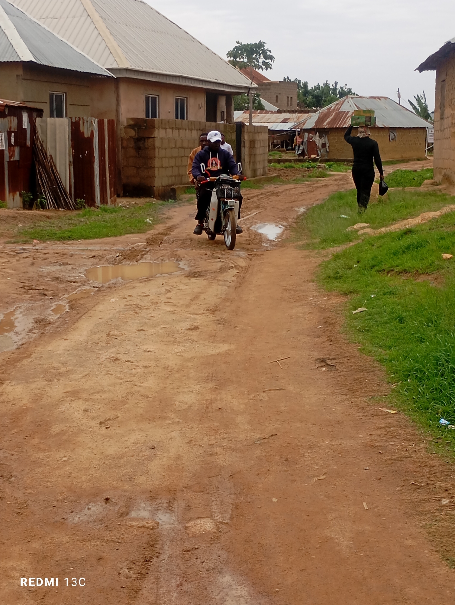 A dirt road in a residential area of Jos, Nigeria, is partially covered in mud and puddles, indicating recent rainfall. Two individuals are on a motorcycle, with one driving and the other seated behind. Another person, dressed in black, walks away from the motorcycle, carrying a black bag in their right hand and a box on their head. Several buildings with corrugated metal roofs are visible on either side of the road. The sky is overcast, suggesting cloudy weather. The scene depicts a common street view in this part of the city.