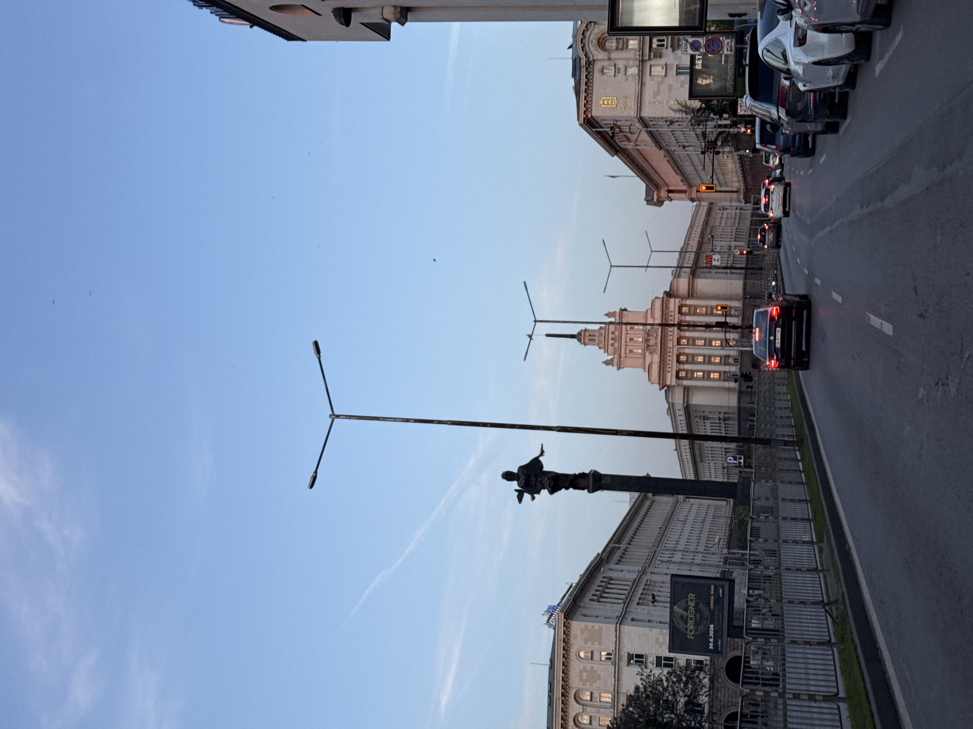 The image, rotated counter-clockwise by 90 degrees, captures a city street scene in Sofia, Bulgaria, at what appears to be dusk or early evening. The sky is a light, clear blue with faint contrails, while streetlights are illuminated and some building windows glow, indicating the transition from day to night. The street surface is visibly wet, suggesting recent rain.
The setting is an urban environment dominated by imposing, large-scale architecture, characteristic of the city center. On the right side of the street, a long line of cars is either parked or stopped in traffic. Several vehicles, mostly dark-colored sedans and SUVs, are visible. Traffic lights with red signals are also present further down the road.
Prominently featured on the left side of the street is a tall, dark monument on a pedestal, which appears to be the Statue of Saint Sophia, a key landmark in Sofia. A distinctive street lamp with a crossbar design stands near the statue. Behind these, large, grand buildings line the street. One particularly notable structure in the mid-ground features a multi-tiered tower or spire, recognized as part of the Largo complex, specifically the former Party House (now part of the National Assembly building). Cyrillic text, "НАРОДНО СЪБРАНИЕ" (National Assembly), is partially visible on this building, confirming its identity.
Metal fences or barriers line the sidewalk, and a large billboard is affixed to them. This billboard displays the text "FOREIGNER" along with a date, "30.4.2026," and smaller text "ALBENA BOGD," possibly advertising an event or concert. Another sign on a building to the right includes the text "HOTEL" and a traffic sign reads "30M" and "BET." The overall scene depicts a typical, albeit grand, city thoroughfare during the evening commute.