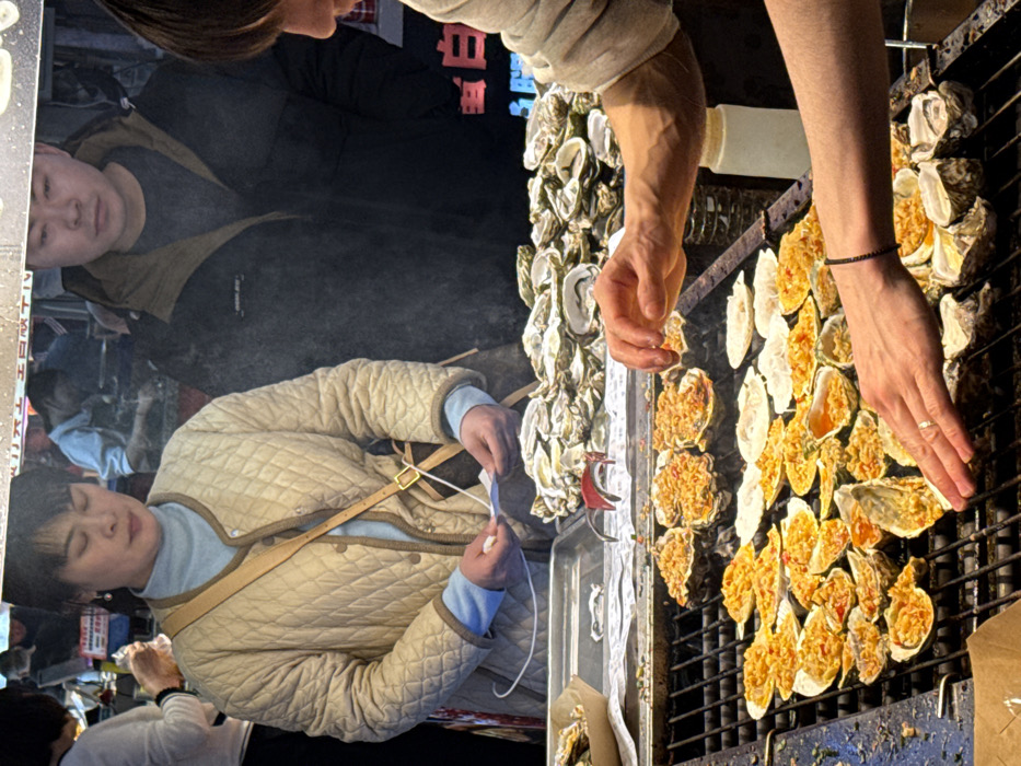 The image depicts a street food stall in Yuzhong District, China, specializing in grilled seafood. In the foreground, a vendor, identifiable by light-skinned forearms, tends to a large metal grill. The vendor's left hand, adorned with a gold ring on the ring finger, is positioned above a half-shell oyster, while the right hand hovers above other grilled items, suggesting a seasoning or manipulation action. A black beaded bracelet is visible on the vendor's right wrist.
The grill is laden with numerous oysters and scallops in their half-shells, topped with a yellowish-orange mixture, actively cooking and emitting steam. To the right of the grill, several raw, unshucked oysters are arranged. Behind the grill, on a metal tray, a substantial pile of open oyster shells, some containing raw oysters, is visible. A white squeeze bottle stands near the right edge of the grill.
Two customers are prominently featured behind the food preparation area. On the left, a female individual with short dark hair wears a light beige quilted jacket over a light blue turtleneck. A tan cross-body strap with a dark brown monogram pattern, possibly from a luxury brand, extends from her left shoulder. She holds a small white paper slip in her hands, appearing to observe the cooking process. To her right, a male individual with short dark hair stands behind her, wearing a dark winter jacket with a brown collar and a dark t-shirt. He also appears to be observing the scene.
The background is dimly lit and blurred, indicating a bustling market environment with indistinct figures and lights, consistent with a night market setting. An overhead translucent covering, possibly plastic or tarp, shows faint white Chinese characters.