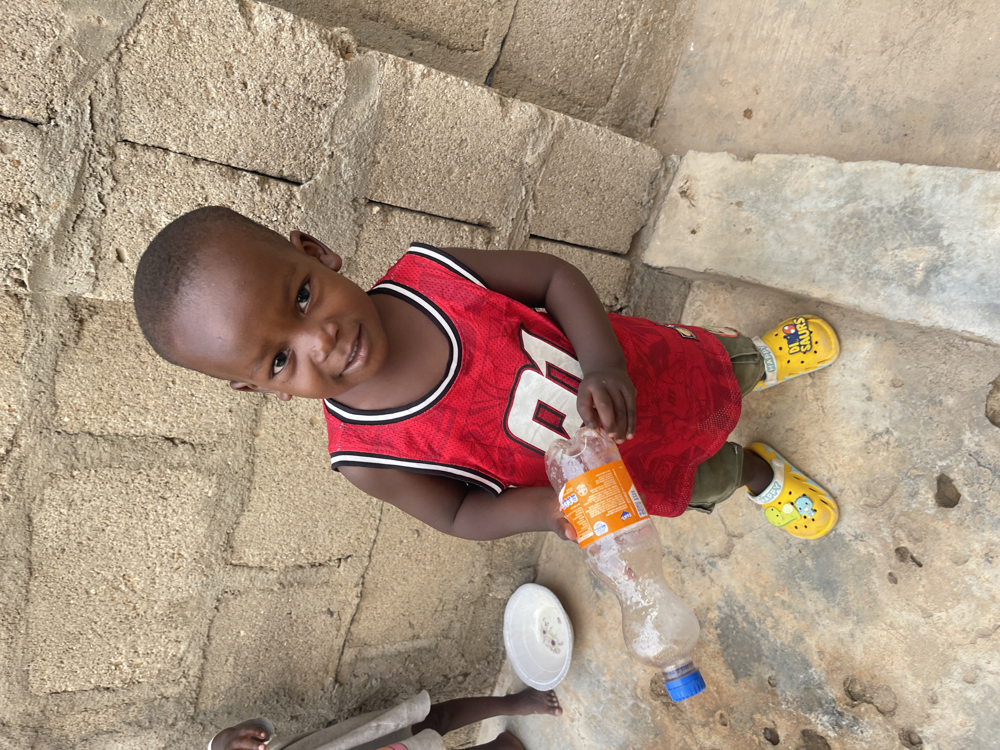 A young child is standing in Unguwar Gabas, Nigeria, holding a plastic bottle. He is wearing a red basketball jersey, green shorts, and yellow sandals. A white bowl is on the ground near him. The background consists of a textured concrete wall and the ground is a mix of concrete and bare earth. Another person is visible from the waist down, wearing light-colored clothing.