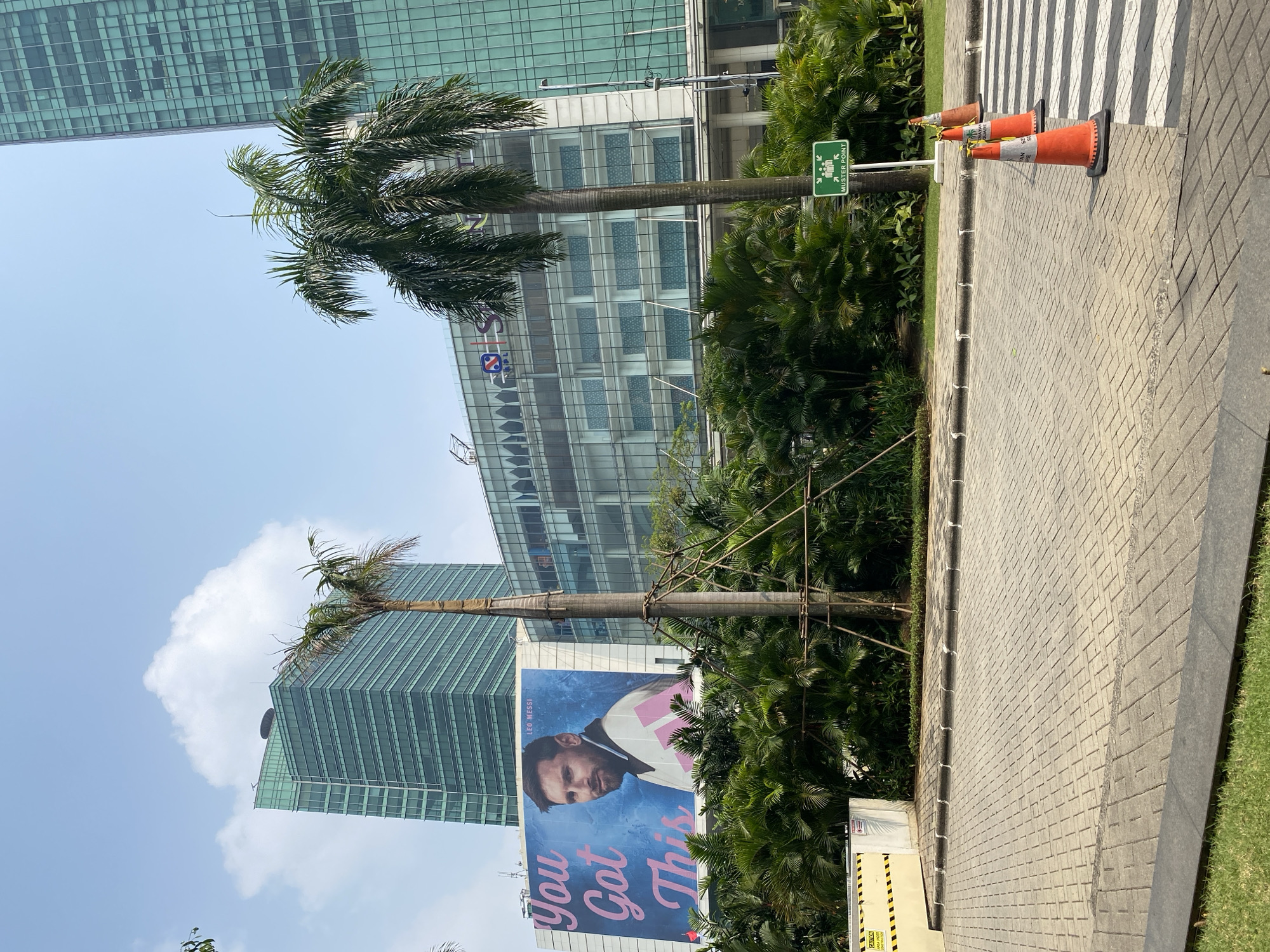 In the city of Central Jakarta, Indonesia, an outdoor scene is depicted with modern architecture and lush tropical vegetation. Dominating the left side of the frame is a large billboard featuring a portrait of soccer player Leo Messi, with the text "You Got This" in pink font against a blue background. To the right of the billboard, a contemporary building with a glass facade stands tall. In the foreground, a paved walkway leads to a crosswalk, with three orange traffic cones placed on the right side of the walkway. Several palm trees are interspersed throughout the scene, adding to the tropical ambiance. A green sign with a pictogram of people and the words "Muster Point" is also visible. The sky is clear blue with a few white clouds.