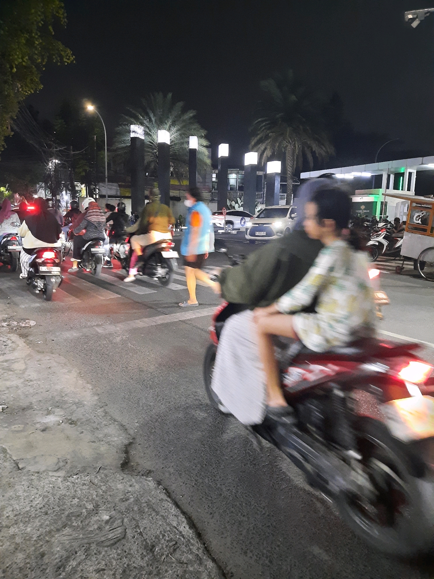 The image depicts a nighttime street scene in East Jakarta, Indonesia, characterized by a busy intersection with several motorbikes and a few cars. The foreground is dominated by a red motorbike with two occupants, their figures blurred by motion. The rider is wearing a patterned shirt, and the passenger, a young individual, is facing away from the camera, also in a patterned shirt. The motorbike is moving through a pedestrian crossing.
In the mid-ground, a group of motorbikes and their riders are also in motion, navigating the intersection. One person is walking across the pedestrian crossing, wearing a blue and orange top and dark shorts. Other motorbikes are visible on the left side of the image, with riders wearing helmets and casual clothing. A few cars are also present, including a white car with its headlights on.
The background reveals urban elements of East Jakarta, including streetlights, palm trees, and modern buildings, one of which appears to be a commercial establishment with illuminated signage. The overall atmosphere is one of nighttime urban activity and traffic flow. The lighting suggests artificial illumination from streetlights, building lights, and vehicle headlights.