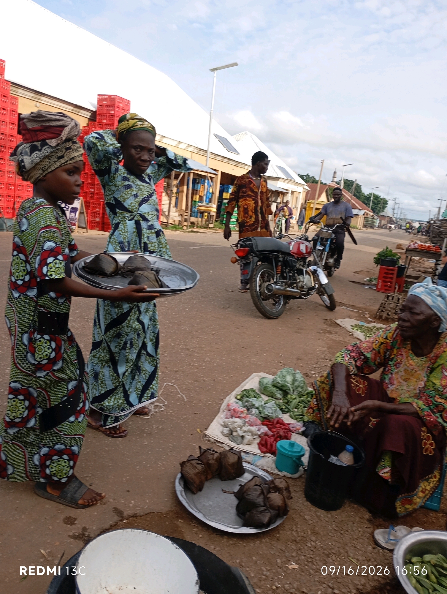 In Shendam, Nigeria, a bustling street market is captured under a cloudy sky. In the foreground, two young girls in vibrantly patterned traditional clothing are featured. The girl on the left, with a headscarf made of layered fabrics, holds a silver tray laden with four wrapped food items, possibly fufu, a staple West African dish. She wears a green, red, and white patterned dress and dark sandals. Next to her, another girl adjusts her headscarf, dressed in a similar green, blue, and white patterned dress.
Behind them, the market buzzes with activity. A man in a brown and orange patterned robe walks away from the camera. A motorcycle is parked in the middle ground, with a man in a blue shirt riding another motorcycle in the background. To the right, an elderly woman, adorned in a colorful, embroidered garment and a blue headscarf, sits on the ground next to a display of fresh produce. A variety of vegetables, including leafy greens and what appear to be peppers, are arranged on a mat. A black bucket containing a bottle and a blue plastic container are also nearby.
The market appears to be situated along a dusty street, with buildings and stalls lining the background. Red crates are stacked prominently to the left, suggesting commerce and inventory. The overall scene depicts a lively everyday marketplace in a Nigerian city.