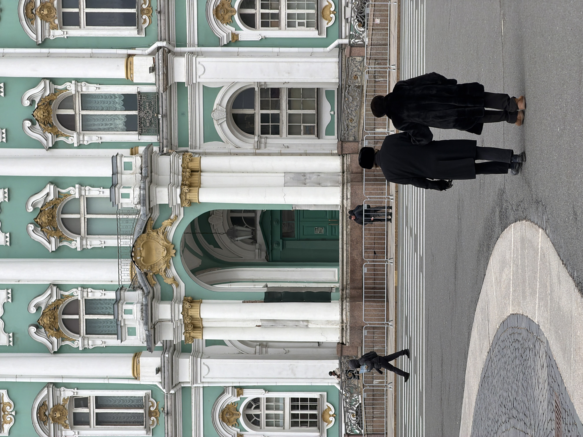 The image depicts a grand, ornate Baroque-style building, likely the Winter Palace in Saint Petersburg, Russia, featuring a pale green facade with prominent white columns and intricate golden decorative elements. The building displays multiple windows of varying shapes, including arched and rectangular, some adorned with gilded frames and iron balconies. A large archway or entrance is visible towards the center of the building's facade.
In the foreground and midground, a wide paved area, possibly a square or street, stretches across the frame. It features asphalt with white painted lines, resembling a pedestrian crossing or designated zone, and a section of decorative dark paving stones forming a semi-circular pattern on the far right. White metal crowd control barriers run along the edge of the building, separating it from the paved area.
Several people are present. In the foreground, with their backs to the viewer, two individuals stand on the paved area, both wearing dark coats and hats; one coat appears to be fur, and the other is a long, dark overcoat. Further back, another person in dark clothing carrying a backpack is seen walking towards the building. Two more smaller figures are visible in the distance near the building's archway.
The scene takes place during the daytime under what appears to be clear or mildly overcast weather, with soft, diffused light illuminating the environment. No visible text is present in the image.