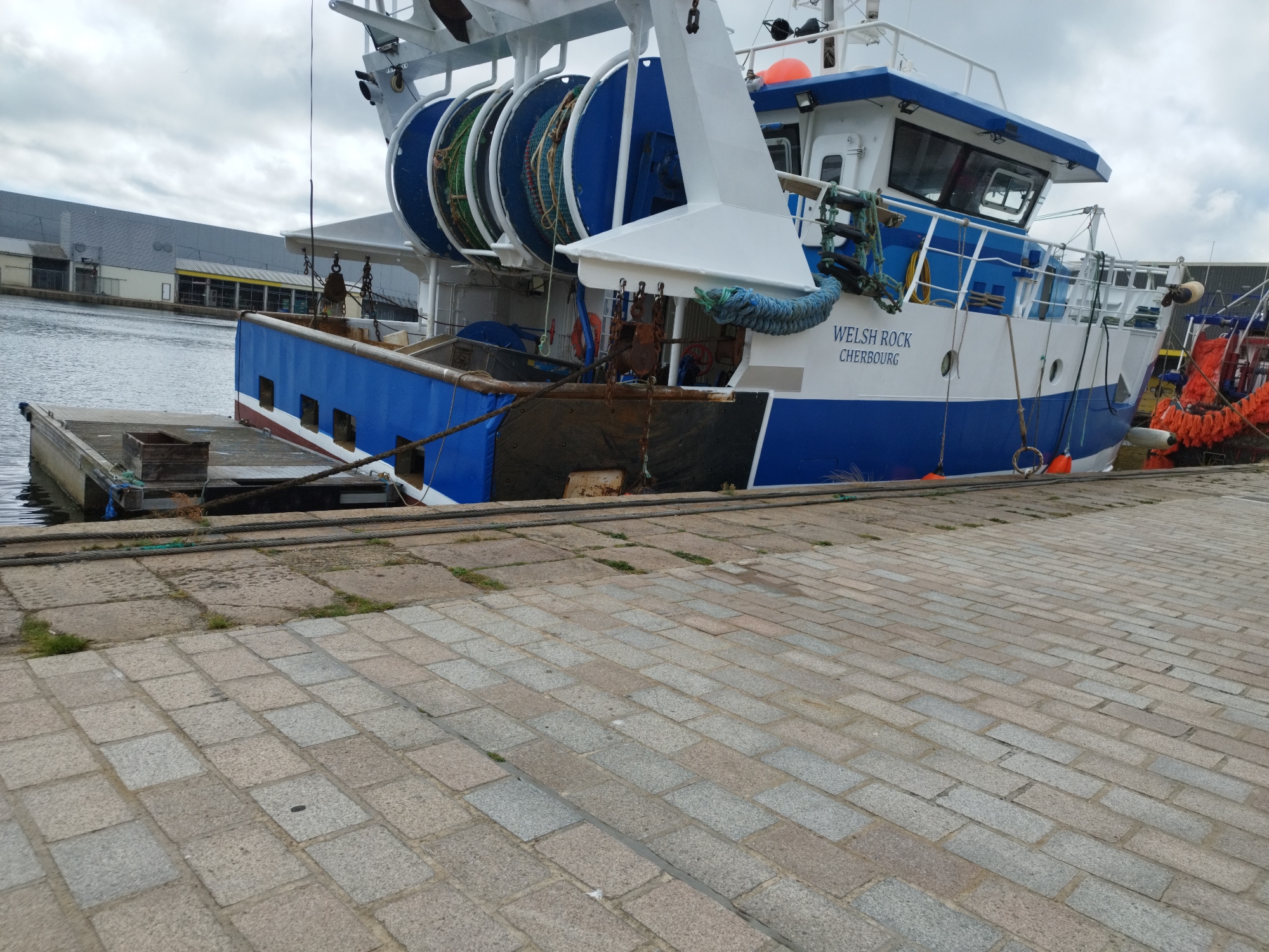 The image depicts the stern section of a blue and white fishing trawler named "WELSH ROCK" docked at a quay in Cherbourg, France. The vessel features large blue spools wound with netting on its deck, a cabin structure, and various ropes and buoys. The quay is paved with granite cobblestones, and a large industrial building with grey cladding is visible in the background across the water. The sky is overcast, suggesting cloudy weather. No people are present in the scene. The scene is static and indicative of a fishing vessel at port.