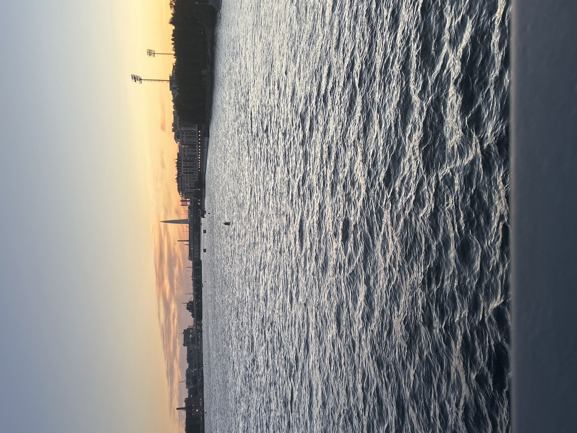 The image depicts a wide body of water with moderate waves, likely a river or bay, in the foreground. The water's surface is dark blue and reflects the twilight sky. In the background, a city skyline is visible against a soft orange and pale blue sky, indicative of either sunrise or sunset. Several buildings, including what appears to be a tall, slender skyscraper, line the distant shore. The cityscape is accompanied by a tree line and what seem to be stadium lights. A red buoy floats on the water's surface. A dark, blurred railing occupies the very bottom of the frame, suggesting the photo was taken from a bridge or elevated viewpoint. The overall scene is calm and serene, with a focus on the natural beauty of the water and sky contrasting with the urban environment.