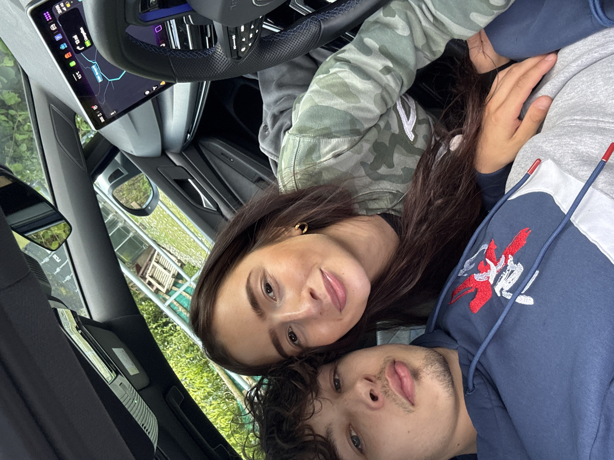 A couple poses in a car for a selfie. The car's navigation screen shows an address in Llangynog, United Kingdom. The woman has long, dark brown hair and is wearing a camouflage jacket. The man has curly, dark brown hair and is wearing a blue and gray hoodie with a red and white logo. They are both looking at the camera and smiling slightly. The car's interior is modern and sleek. Outside the window, greenery is visible.