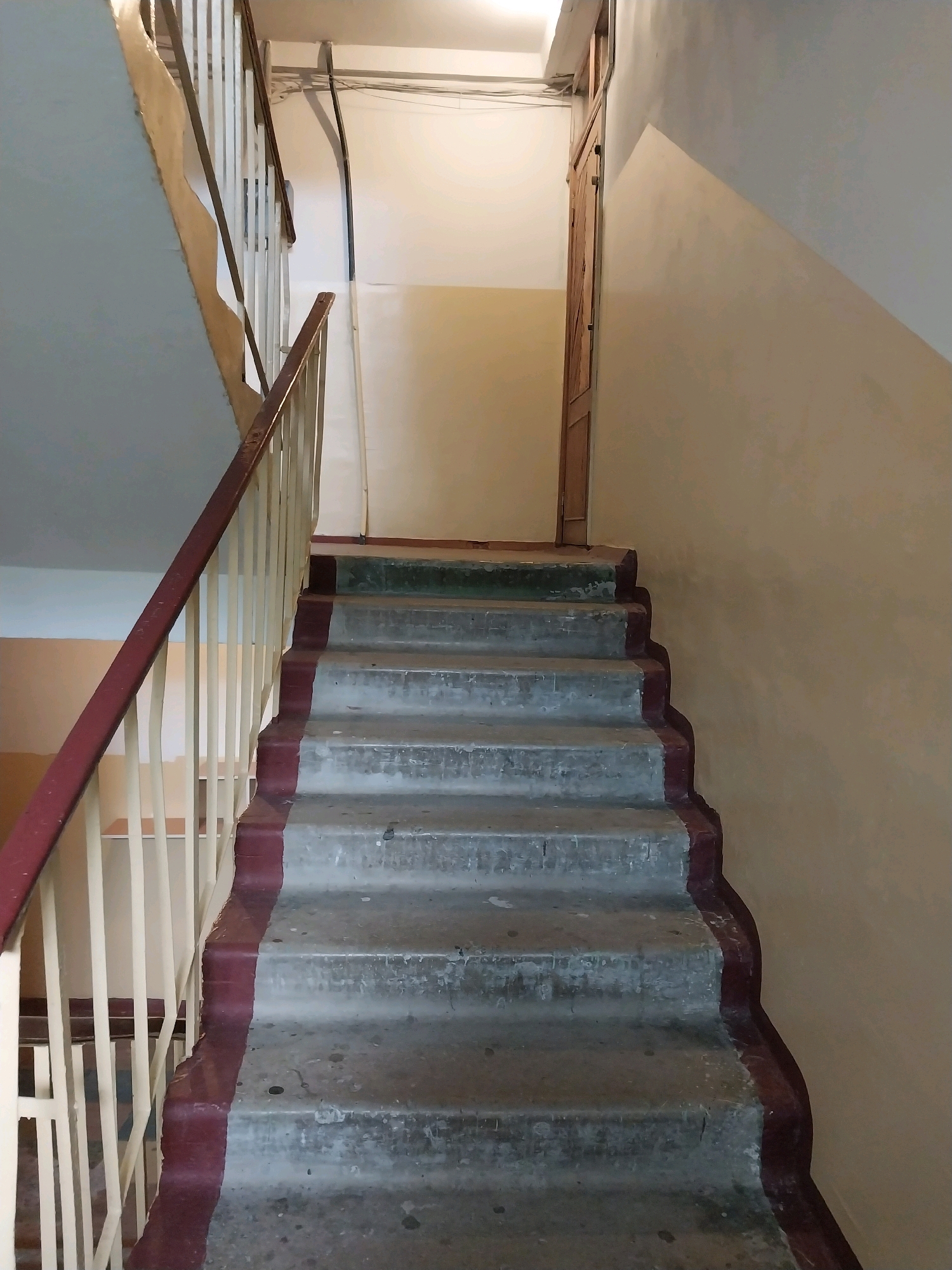 This is a medium-shot, eye-level, vertical photograph of a concrete staircase in an apartment building in Dnipro, Ukraine. The staircase has seven visible steps with a dark red painted trim along the edge of each step. The railing is made of light-colored vertical bars with a dark red wooden handrail. The walls are painted in a light yellow and beige color. A wooden door is visible at the top of the stairs. Electrical conduits and wires run along the ceiling. The scene depicts an interior common area of an apartment building.