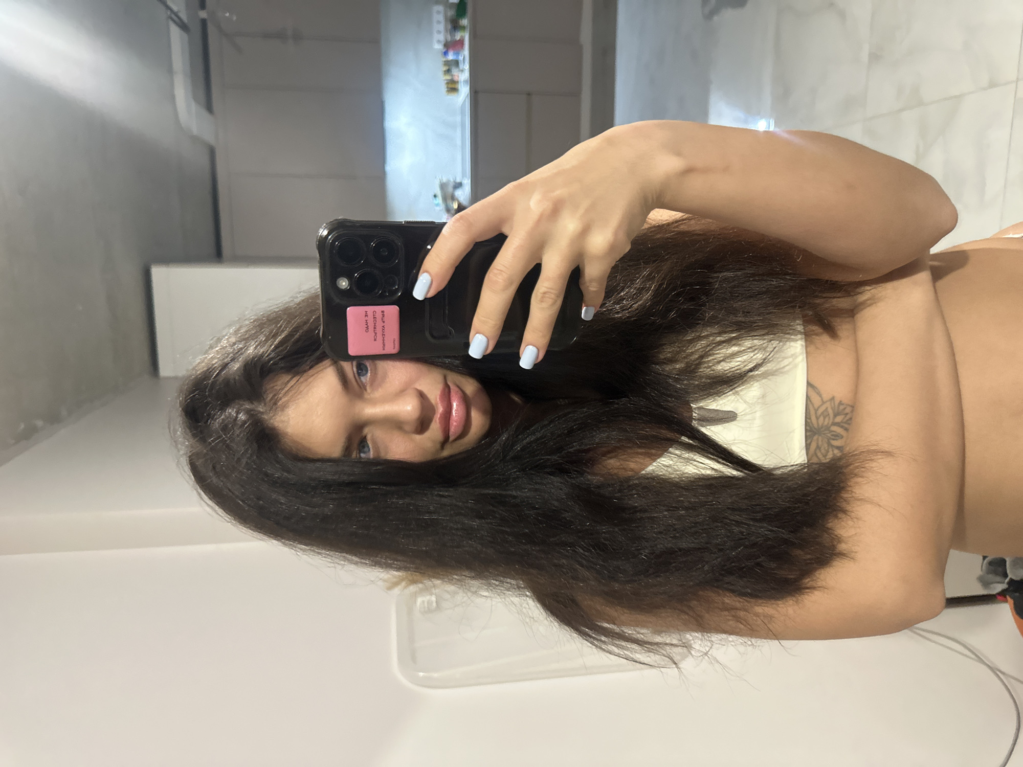 A young woman is depicted taking a mirror selfie indoors, likely in a private residence in Khimki, Russia. She has long, dark hair and striking blue eyes, with her lips slightly parted in a neutral to soft expression. She is wearing a light-colored (possibly white or beige) top. A tattoo with a floral design is visible on her upper right arm/shoulder area. Her fingernails are painted a light blue color. She holds a dark-colored smartphone, which appears to be an iPhone, in her right hand, positioned to capture her reflection. A pink sticker or insert is visible on the back of the phone case. This sticker displays reversed, partially legible Cyrillic text due to the mirror reflection. The background, also visible in the mirror, consists of modern, minimalist interior elements, including white tiled walls and what appears to be a counter or shelf. The overall setting suggests a bathroom or a similar utility area within a home.