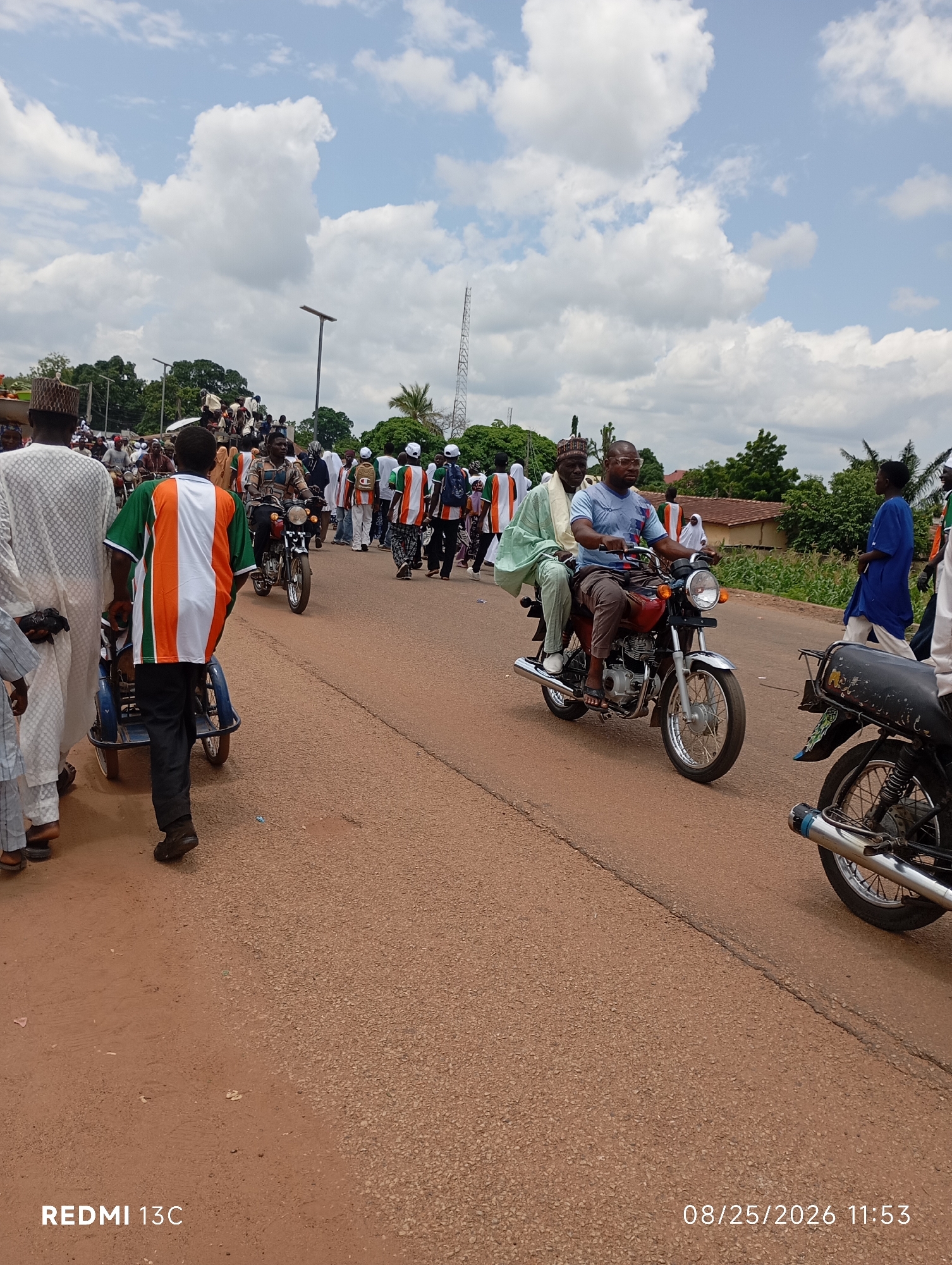In the city of Shendam, Nigeria, a street scene unfolds under a cloudy sky, with a crowd of people gathered and moving along a paved road. The atmosphere suggests a public event or procession.
In the foreground, a man in traditional white attire stands to the left, looking towards the side. Next to him, another man in a vibrant orange, green, and white striped shirt walks, pushing a small blue cart with two wheels. Further down the road, two men ride a red motorcycle. The driver wears a blue t-shirt and glasses, while the passenger is in a light green robe. Both appear to be looking forward.
In the middle ground, several other individuals are visible, some riding motorcycles and others walking. Many are wearing clothing in similar colors to the man in the foreground, suggesting a common affiliation or theme. The background shows a line of buildings, trees, and a telecommunications tower, indicative of a semi-urban environment. The road is an asphalt surface with some texture.
The date and time stamp at the bottom right corner of the image reads "08/25/2026 11:53," and the text "REDMI 13C" is at the bottom left.