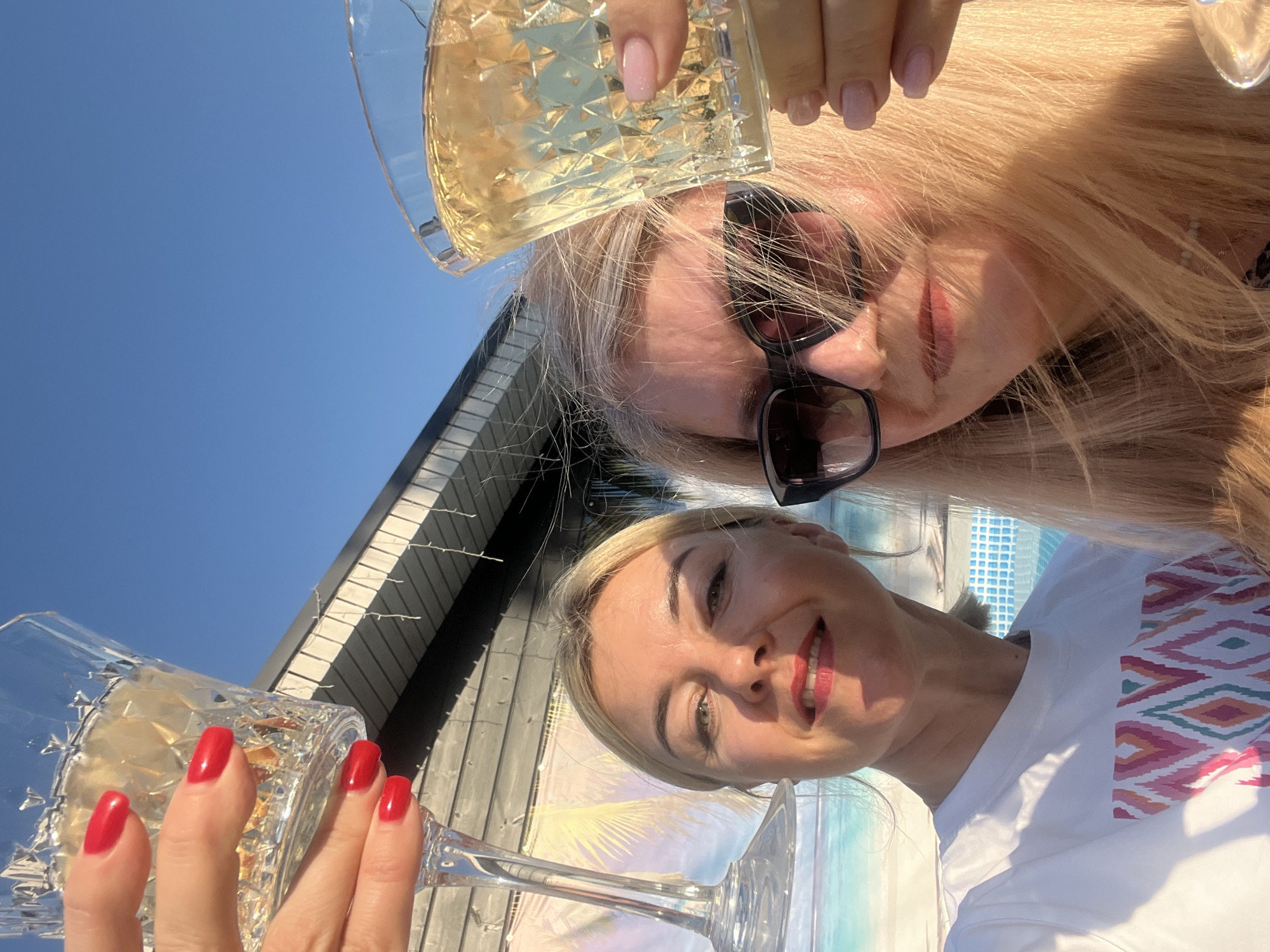Two women are raising glasses of a light-colored beverage, possibly champagne or white wine, in a toast. The woman in the foreground has blonde hair, fair skin, and is wearing a white t-shirt with a colorful geometric pattern. Her nails are painted red. The woman in the background has lighter blonde hair, is wearing sunglasses, and a light-colored manicure. They are both smiling. The backdrop includes a blue sky, the roof of a building with vertical siding, and a light-colored structure with a patterned surface. The exact location in Новая Чернушка, Russia is not determinable without further information.