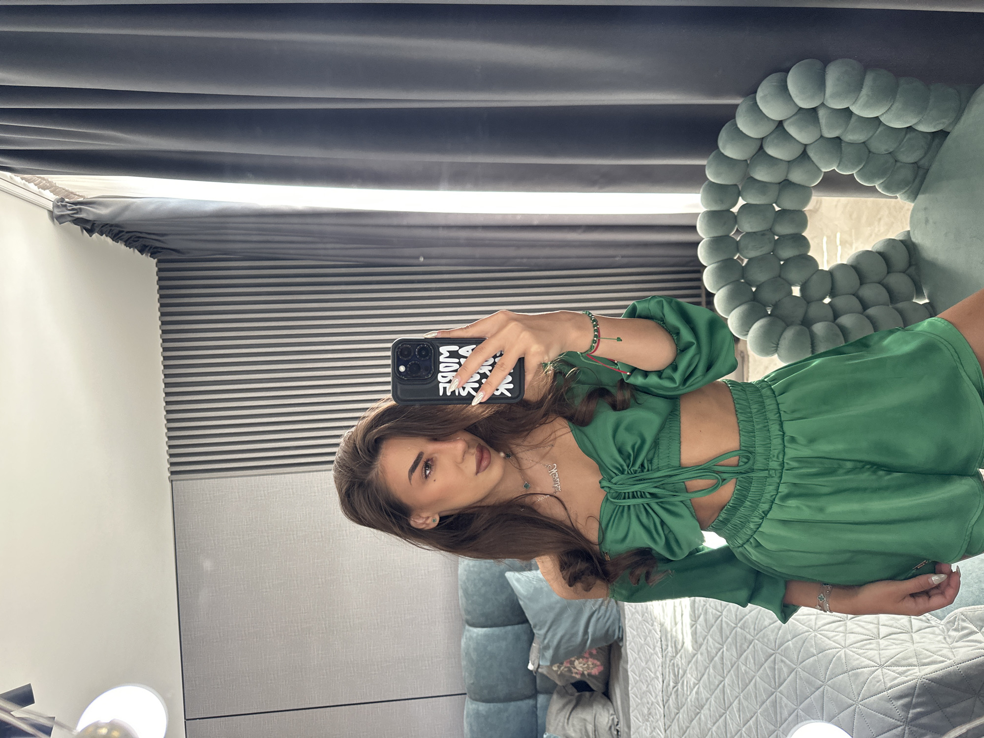 A young woman with long, dark brown hair is taking a selfie in a bedroom. She is wearing a green satin two-piece outfit consisting of a ruched crop top with off-the-shoulder sleeves and matching shorts. She has on a delicate necklace with a pendant and several bracelets on her left wrist. The woman is holding a black smartphone in her right hand, with a phone case that has white text on it.
The background of the image shows a bedroom with a large, quilted headboard on a bed. To the left of the bed is a floor lamp with a circular white shade. Behind the woman, on the wall, is a textured gray paneling with vertical stripes. To the right, dark gray curtains are drawn, with a bright light filtering through the gap between them. In the foreground, on the right side of the image, is a plush, light green armchair with a distinctive rounded, bubble-like design.
The style of the room suggests a modern, luxurious interior. The overall impression is one of a well-decorated personal space. There are no indications that this scene is specifically located in Sofia, Bulgaria.