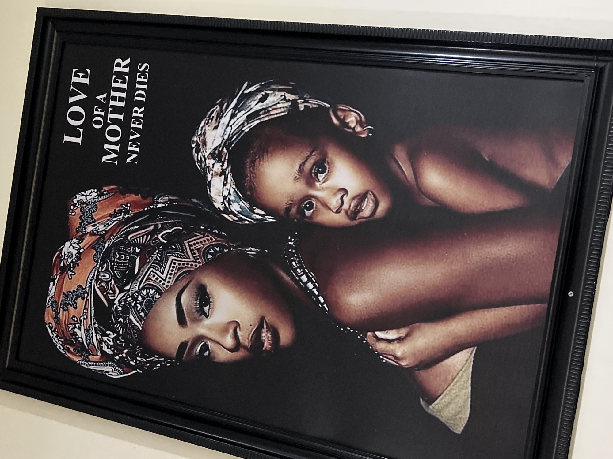 A framed picture, angled slightly to the right, depicts a portrait of a woman and a child against a dark, possibly black, background. The frame itself is black with intricate, ridged detailing along its inner and outer edges.
In the foreground, a woman with dark skin wears a vibrant, patterned headwrap featuring shades of orange, brown, black, and white. She looks towards the viewer with a serene expression, a faint smile, and appears to have eye makeup. Her hand is visible near the bottom right, and a beaded necklace-like element drapes across her shoulder.
Behind and partially to the left of the woman, a young child with dark skin is visible, looking directly at the viewer. The child also wears a patterned headwrap, distinct from the woman's, with patterns in grey, white, and black. The child has wide, curious eyes and a slightly open mouth. The overall composition suggests a close, loving bond between the two, likely a mother and child.
On the left side of the artwork, white capitalized text is prominently displayed, reading: "LOVE OF A MOTHER NEVER DIES". "LOVE" and "MOTHER" are in a larger font size than "OF A" and "NEVER DIES".
The image captures a moment of maternal affection and cultural identity, potentially in a home or gallery setting in Gwarinpa, Nigeria, given the provided context. The warm skin tones and traditional headwraps hint at an African cultural context.