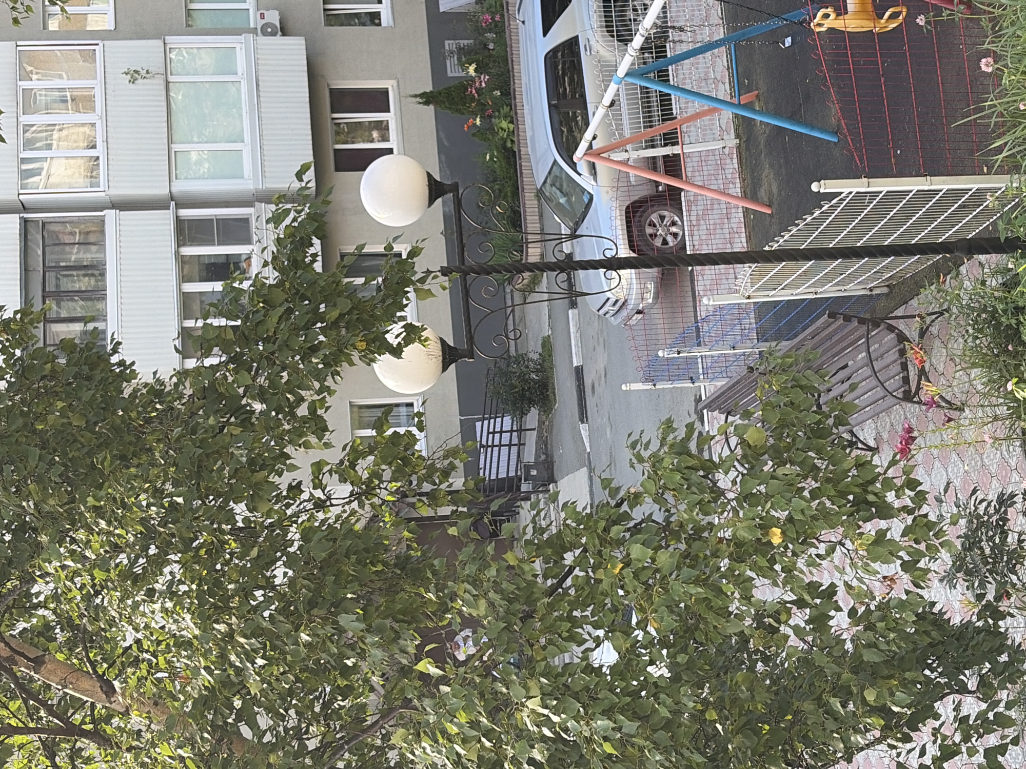 The image captures a view of a courtyard in Korsakov, Russia. A large tree with green leaves dominates the left side of the frame, its branches partially obscuring a multi-story apartment building with light-colored facade and numerous windows. In the foreground, a playground with a swing set, partially visible, is enclosed by a fence. A silver SUV is parked on the asphalt surface in front of the building. Two spherical outdoor lamps are mounted on a decorative metal post to the right of the tree. A wooden bench is situated near the playground fence. In the background, an entrance to the building is visible, with a ramp leading up to it, and some flowering plants are present in front of the building. The ground surface is a mix of asphalt and paving stones.