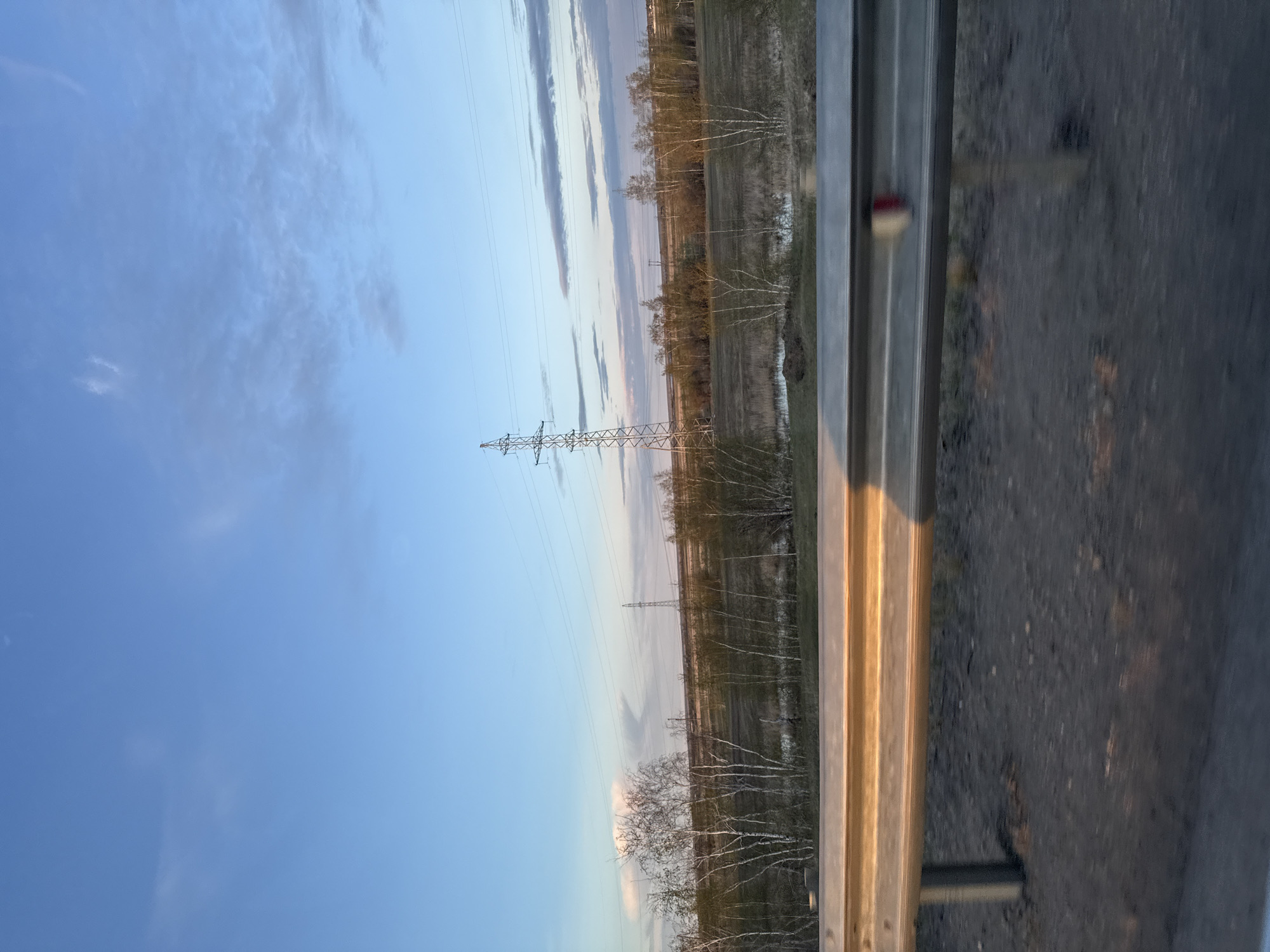 The image shows a landscape with a transmission tower in the distance. The sky is blue with some clouds. There are trees and fields, and what appears to be a ditch or small body of water. In the foreground, there is a metal guardrail and asphalt, suggesting the view is from a road. The location is implied to be Syrdakh, Russia, but there are no details in the image to confirm this. There are no people visible.