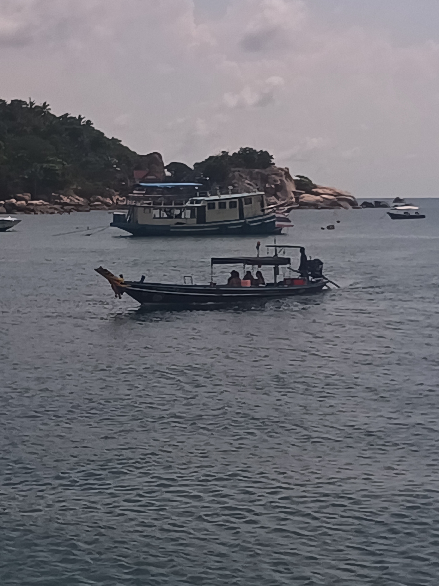 The image shows a coastal scene in Tambon Ko Tao, Thailand. The foreground is dominated by the rippled, grey ocean water. In the mid-ground, a longtail boat with several passengers moves across the water, powered by an outboard motor. A decorative figurehead adorns the bow of the boat. Further back, a larger boat is anchored. In the background, a lush, green, rocky island rises from the sea. The sky is overcast, casting a muted light over the scene.