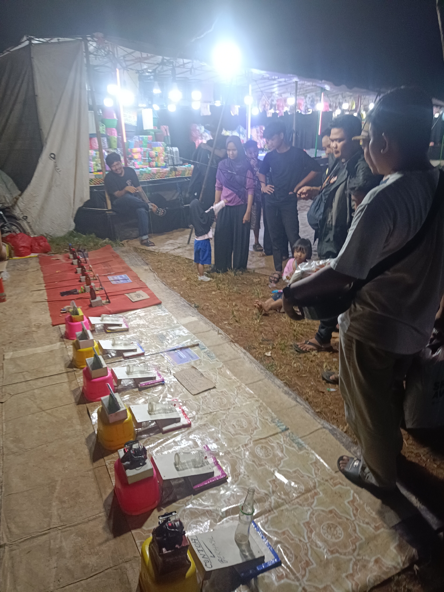 The image depicts an outdoor scene at night, likely a market or a temporary event. Several people are present, including adults and at least one young child. In the foreground, a row of objects is arranged on mats, suggesting a game or a stall. These objects appear to be small bottles, possibly part of a game of chance or skill. The lighting is artificial, with bright overhead lights illuminating the area, creating harsh shadows.
The setting includes what looks like a stall or tent in the background, with stacked colorful items, possibly goods for sale. The ground is a mix of dirt and covered areas with patterned mats. The overall atmosphere seems casual and communal, with people gathering and interacting. The time of day is clearly night, indicated by the artificial lighting and dark sky.
The individuals in the image are dressed casually. Some are standing and observing, while others are seated or engaged in conversation. One person in the foreground is in focus, wearing a light-colored t-shirt and dark pants, with a bag slung across their shoulder. A young child in a pink shirt and blue shorts is also visible, looking towards the camera.
The activity appears to be a nighttime gathering at a market or fair, possibly involving a game or activity set up in the foreground. The mood seems relaxed and social. There is some visible text on a white sheet on the mat in the bottom right corner, which appears to read "CONTOH BERHASIL" in Indonesian, translating to "SUCCESSFUL EXAMPLE". This text, along with the arrangement of items, further suggests a game or a demonstration. The location context of Depok, Indonesia, implies this is likely a local event.