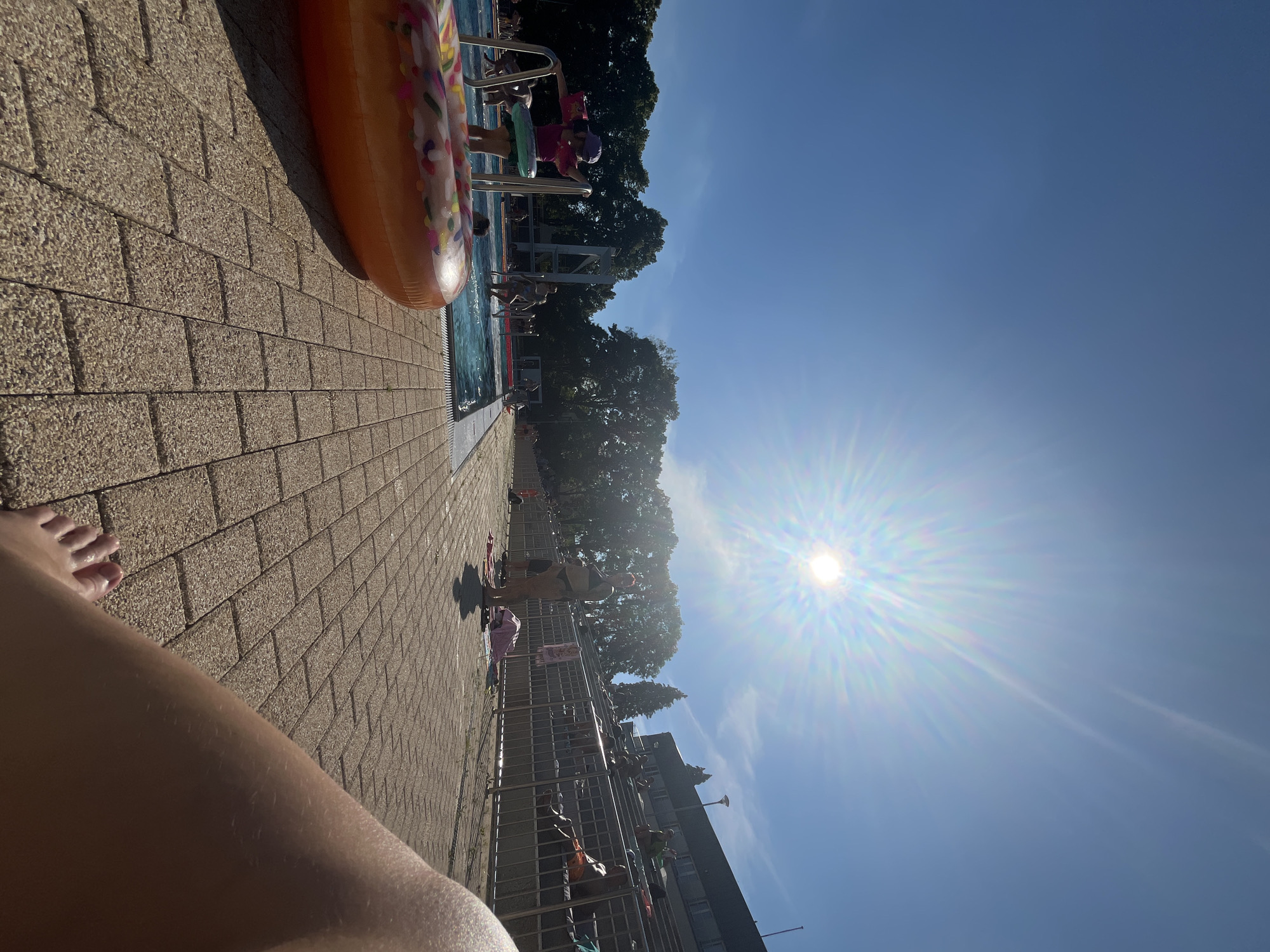 The image depicts an outdoor swimming pool on a sunny day. The sun is bright and high in the sky, casting a strong glare. In the foreground, a portion of a person's leg and foot is visible, resting on a patterned brick patio. To the left, an inflatable ring shaped like a donut is partially visible on the patio.
The middle ground features the swimming pool with several people swimming and standing in and around it. A young child in a pink swimsuit and purple hat is holding onto the pool ladder, with an inflatable ring around their waist. Further back, more people are seen in the pool and around its edges. Beyond the pool, a metal fence separates the pool area from a tiered seating area.
In the background, a building with multiple windows is visible, and beyond that, a line of trees. The overall scene conveys a relaxed atmosphere, likely at a public pool or resort, on a warm, clear day. The location is not specified beyond being outdoors in an urban or suburban setting.