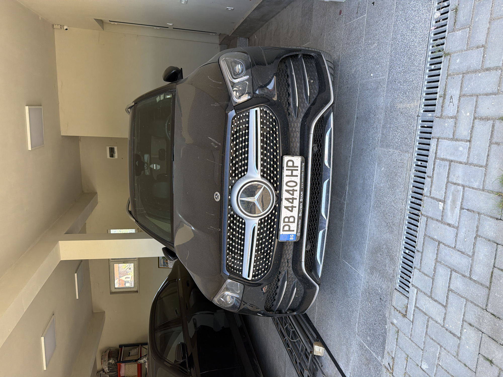 The image depicts a black Mercedes-Benz SUV parked in what appears to be a garage or covered parking area. The front of the vehicle is prominently displayed, including the license plate, which reads "PB 4440 HP" and indicates a Bulgarian registration. The car is somewhat dusty, suggesting it has not been recently cleaned. The background shows a paved area with a drainage grate.
Based on the license plate and publicly available information, specifically the PB prefix, the car is registered in the region of Pazardzhik, Bulgaria, not Asenovgrad. The setting is a garage or covered parking spot, indicated by the surrounding walls and structure. The lighting suggests the space is partially enclosed. No people are visible in the frame.