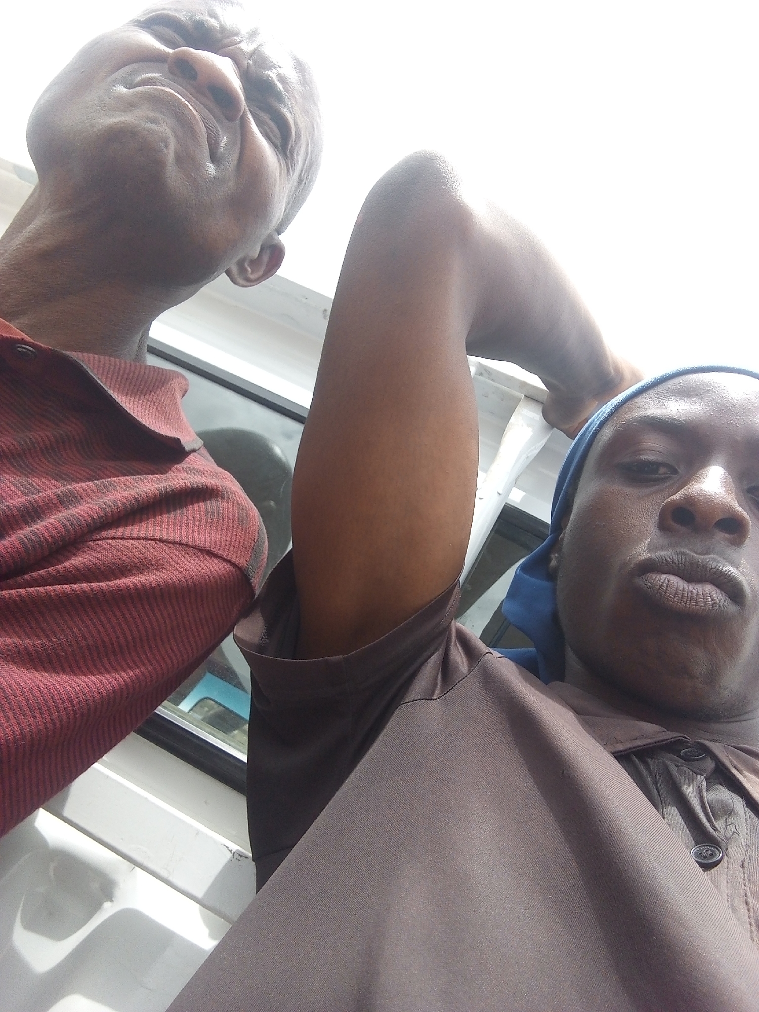 Two individuals, likely adult males, are captured in a low-angle, close-up selfie, possibly from within a vehicle or enclosed space. The person in the foreground, facing the camera, wears a blue head covering and a dark brown collared shirt with visible buttons. Their arm is raised, and their head is tilted back. The second individual is partially visible in the upper left of the frame, also looking upwards. They are wearing a horizontally striped maroon and black collared shirt. The background appears to be a bright, possibly overcast sky, with glimpses of what might be a vehicle's interior, including white paneling and a reflective surface that suggests a window. The scene's location in Jos, Nigeria, is not visually evident from the provided image and would require external information.