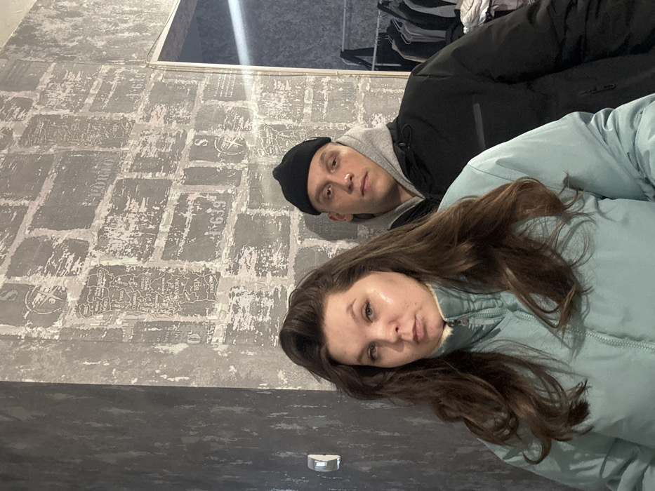 The image depicts two individuals, one female and one male, posing indoors against a textured wall. The female, positioned in the foreground and slightly to the left, has long, dark brown hair and wears a light blue puffer jacket with a cream-colored fleece collar. Her facial expression is neutral, with a slight pout. The male stands slightly behind her, to the right. He wears a black beanie, a light grey hooded sweatshirt, and a black puffer jacket. His gaze is directed slightly off-camera to the right, and his expression is neutral.
The background wall is covered with a patterned wallpaper designed to resemble distressed grey brickwork. This wallpaper features various graphical elements, including a detailed map of Portugal with city names such as Porto, Viseu, Coimbra, Évora, Beja, and Faro. Other design elements include the word "PORTO," numbers like "639," and compass rose symbols. To the far right, a clothes rack draped with various garments is partially visible. A wall-mounted light switch or electrical outlet is present on the left side of the frame. The indoor lighting appears artificial. The visual content of the image does not provide identifiable features to confirm a specific location within Perm, Russia.