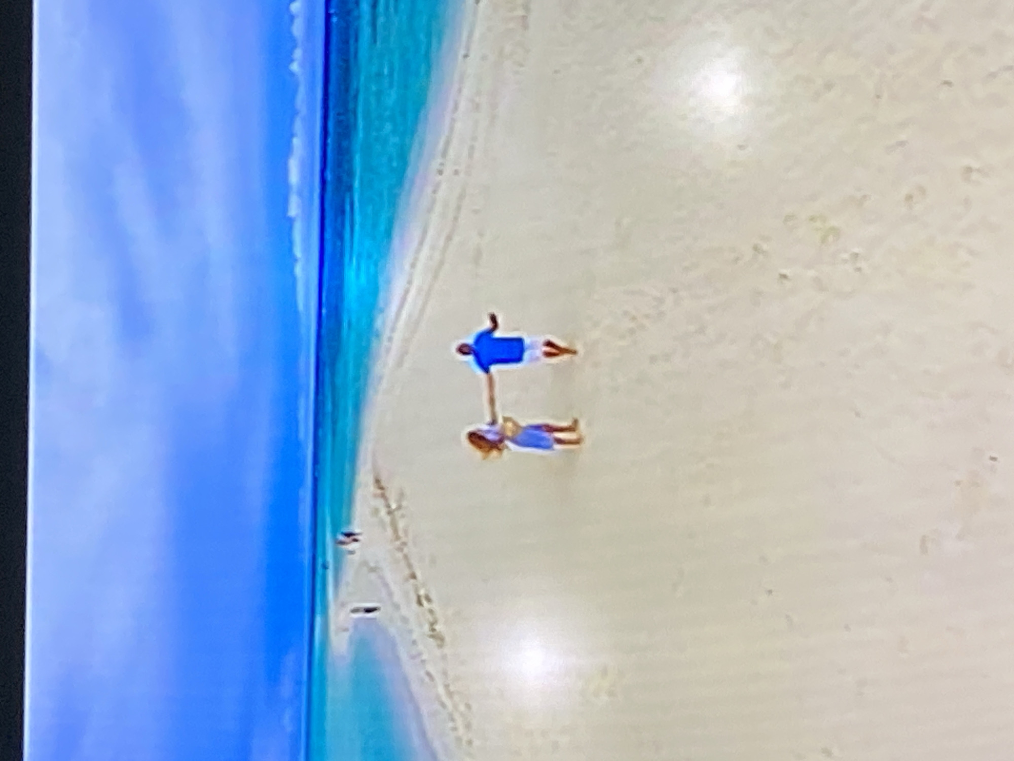 The image depicts a serene beach scene with two individuals holding hands and walking on a sandbar. The person on the left is a woman with long, brown hair, wearing a blue dress. She is holding hands with the person on the right, a man wearing a blue polo shirt and white shorts. They are walking towards the right side of the frame. In the background, there are other figures walking further down the sandbar and a stretch of turquoise water. The sky above is a gradient of blue, with some lighter clouds visible. The scene appears to be a tropical or subtropical location. This image does not depict any location in the city of Yekaterinburg, Russia.