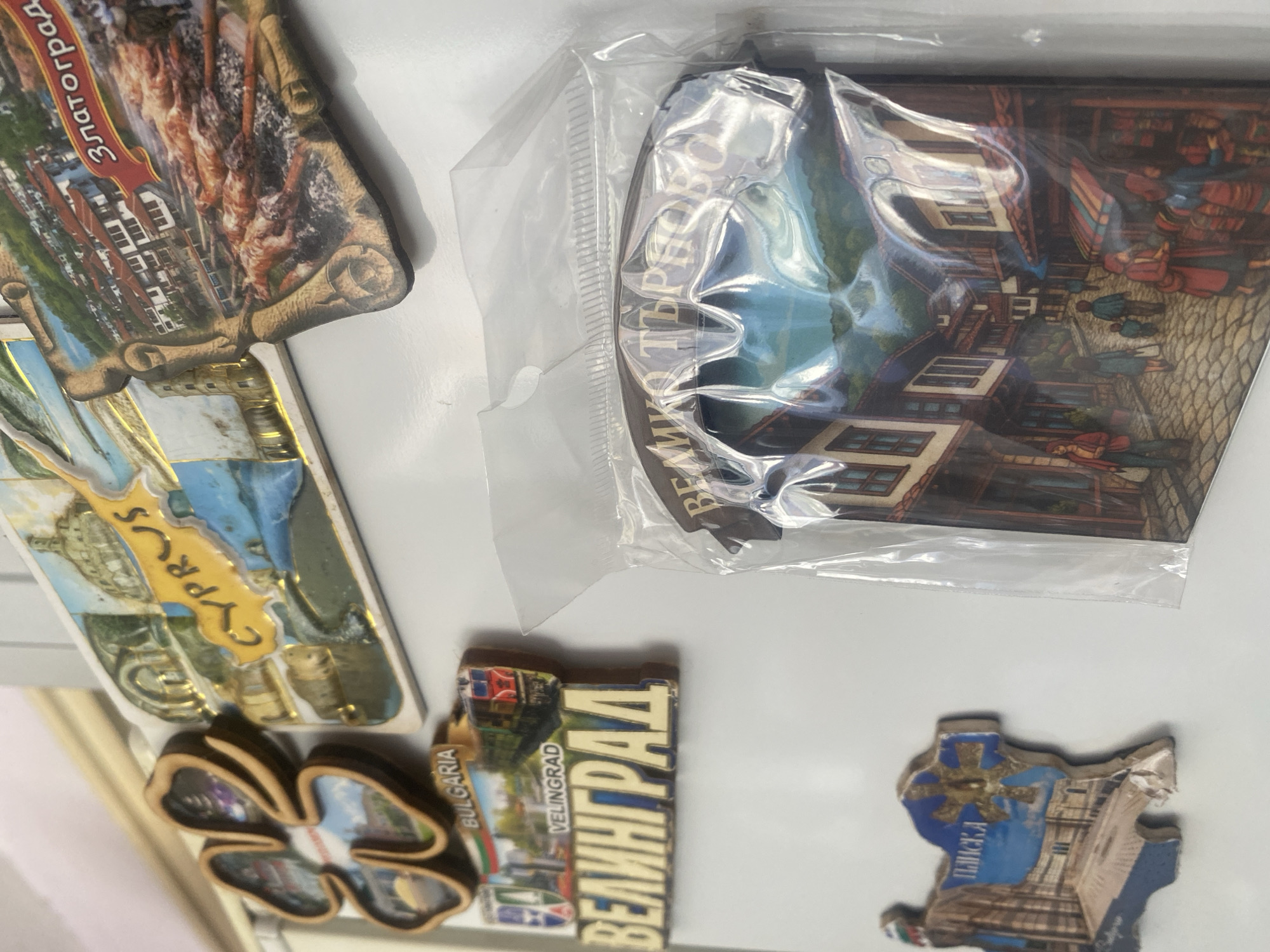 The image shows a collection of souvenir refrigerator magnets against a white background. From left to right, the magnets depict:
1. A scene of Zlatograd, Bulgaria.
2. A scene from Cyprus.
3. A clover shape
4. A scene of Velingrad, Bulgaria.
5. A scene of Veliko Tarnovo, Bulgaria, still in a plastic bag.
6. A scene of Pliska, Bulgaria.
There are no people visible in the image except for the people depicted on the magnets.
The city of Ivaylovgrad, Bulgaria is not directly depicted or mentioned on any of the souvenirs.