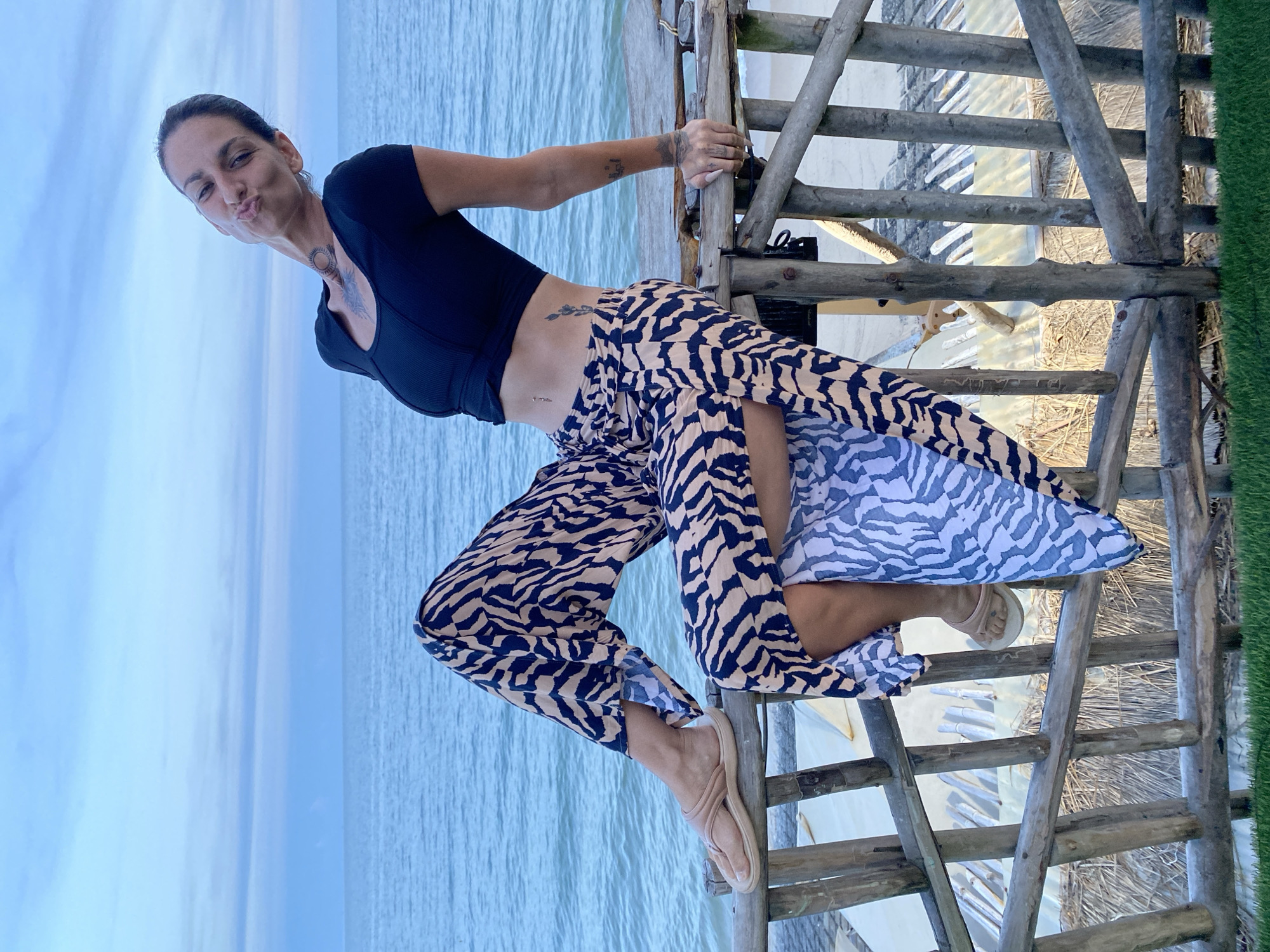A woman with dark hair and tattoos is sitting on a wooden railing overlooking the ocean. She is wearing a black crop top and a flowing skirt with a black and beige zebra print. Her legs are crossed, and her right foot is resting on the railing. The woman is making a kissing face and tilting her head back. The ocean is blue and calm, and the sky is overcast. The background shows a sandy beach and some thatched-roof structures. The scene appears to be a casual outdoor setting, possibly a resort or a beachside cafe. The woman's attire and the tropical environment suggest a warm climate. The overall mood of the image is playful and relaxed. The exact location in Phu Thuy Ward, Vietnam, is not specified by visual cues in the image.