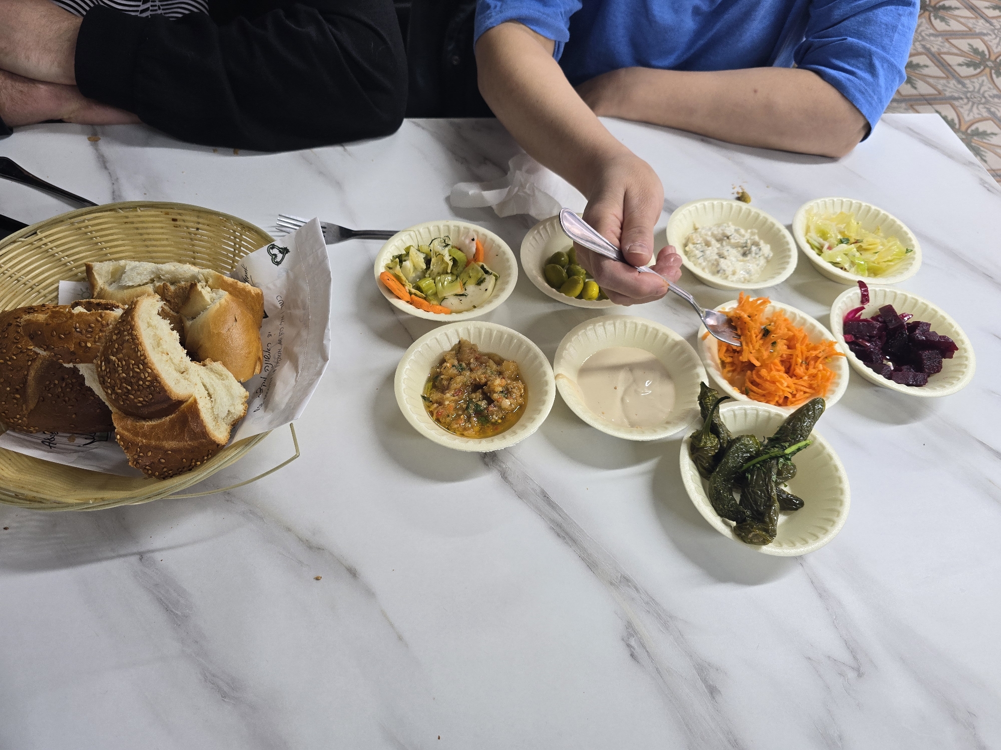 A medium eye-level shot shows two individuals sitting at a white marble-patterned table in Netivot, Israel. One individual, wearing a black shirt, has their arms and hands visible. The other individual is wearing a blue shirt and is holding a silver fork, preparing to eat. On the table, there is a basket of bread, various small white dishes containing pickles, carrots, cabbage, beets, olives, tahini sauce, a spicy tomato mixture, and green peppers. There are two forks on the table to the left of the frame.