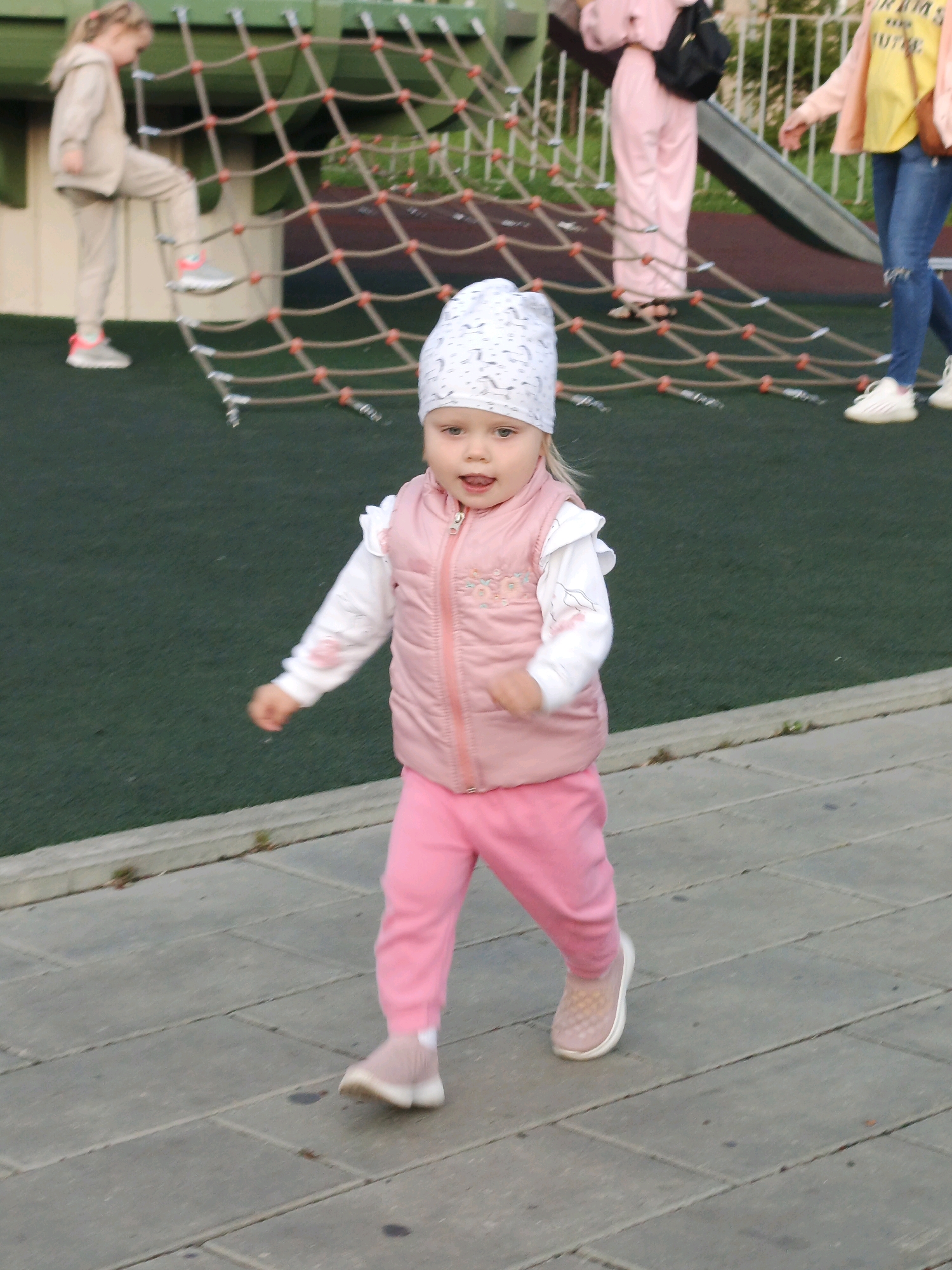 In Zlatoust, Russia, on a paved area adjacent to a playground, a young girl in pink leggings, a white long-sleeved shirt with pink trim, and a pink puffer vest is walking towards the viewer. She is wearing a white patterned hat and light pink slip-on shoes. Her mouth is open, and she appears to be mid-stride with her left arm extended slightly forward. In the background, a rope climbing structure for children is visible, with another child in a beige outfit ascending it. Further back, other individuals are present near a slide and playground equipment. The ground is a dark green rubberized surface common in playgrounds, transitioning to the lighter grey paving stones where the girl is walking. The overall scene depicts a day at a public park.