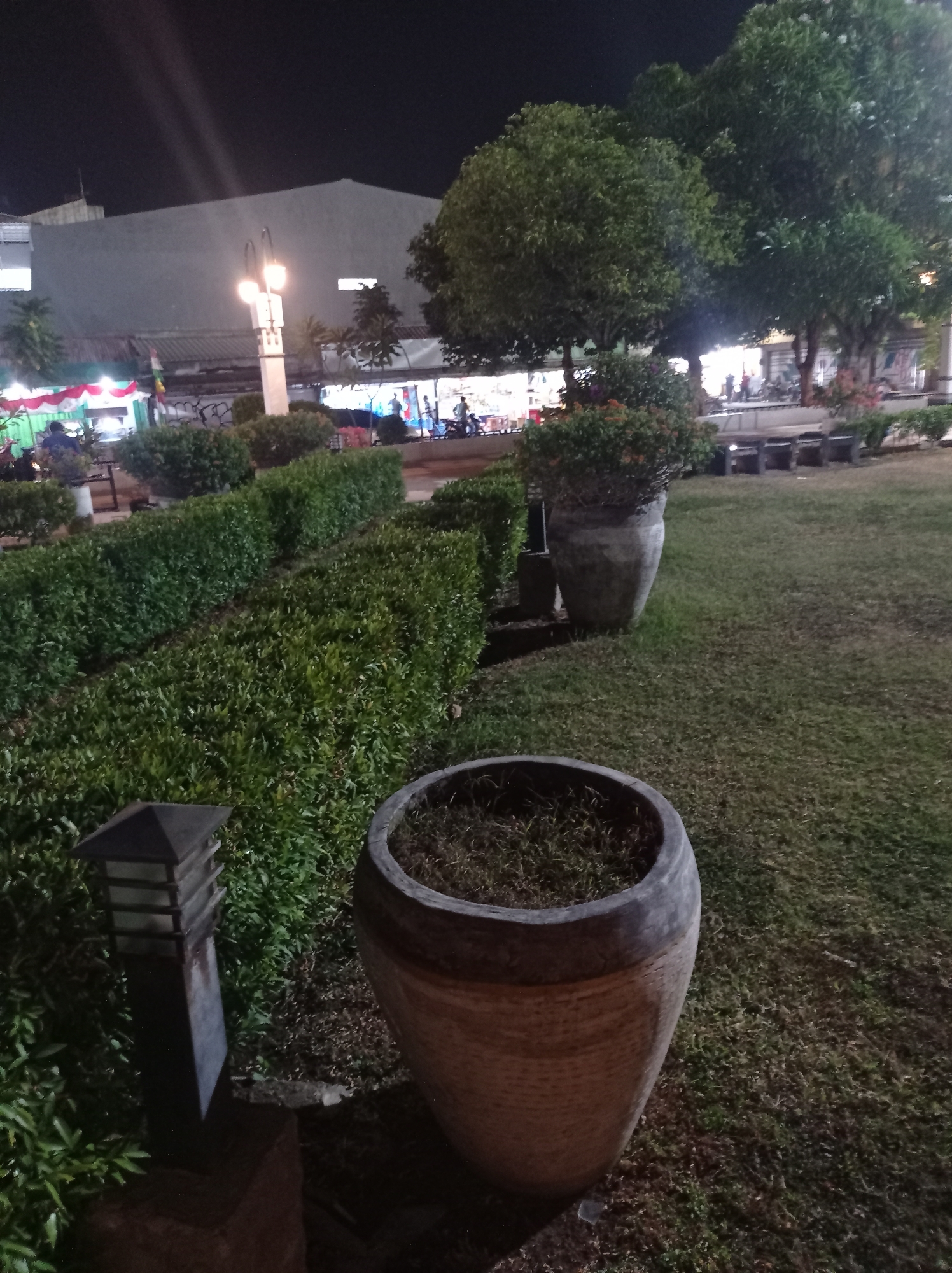 The image depicts an outdoor scene at night, likely in a public area within Tegal, Indonesia. In the foreground, a large, textured ceramic planter filled with dry grass sits on a grassy area. To the left of this planter, a dark, rectangular, tiered garden light is positioned on a dark surface. Further back, a row of neatly trimmed green bushes forms a hedge, with a large, gray, urn-shaped planter containing a green, leafy plant positioned behind it. The background features more trees and some buildings, one of which has a grey facade. In the distance, there are indications of human activity, with several people visible near stalls or shops that are illuminated by colorful lights. Benches are also visible in the midground. The overall impression is a well-maintained park or public square at night.