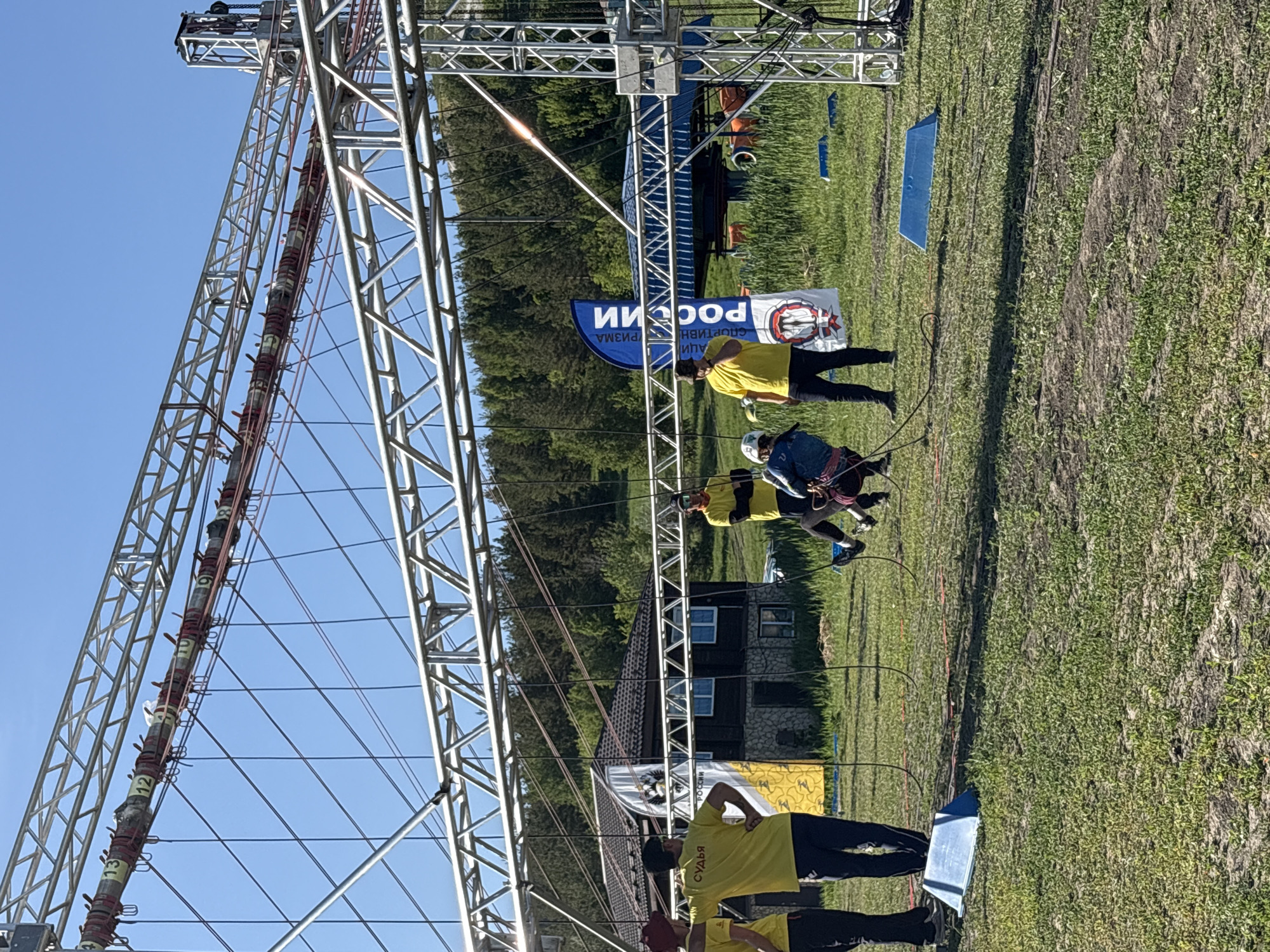 The image depicts an outdoor obstacle course in Khvalynsk, Russia, featuring a tall, metal structure with numbered markers. Two people are navigating a high wire element of the course, while individuals in yellow shirts, labeled "СУДЬЯ" (Judge) appear to be monitoring them from the ground. A banner with "РОССИИ" (Russia) is visible nearby. The environment includes grassy terrain, trees, a building with visible windows and roof. The sky is clear and blue.