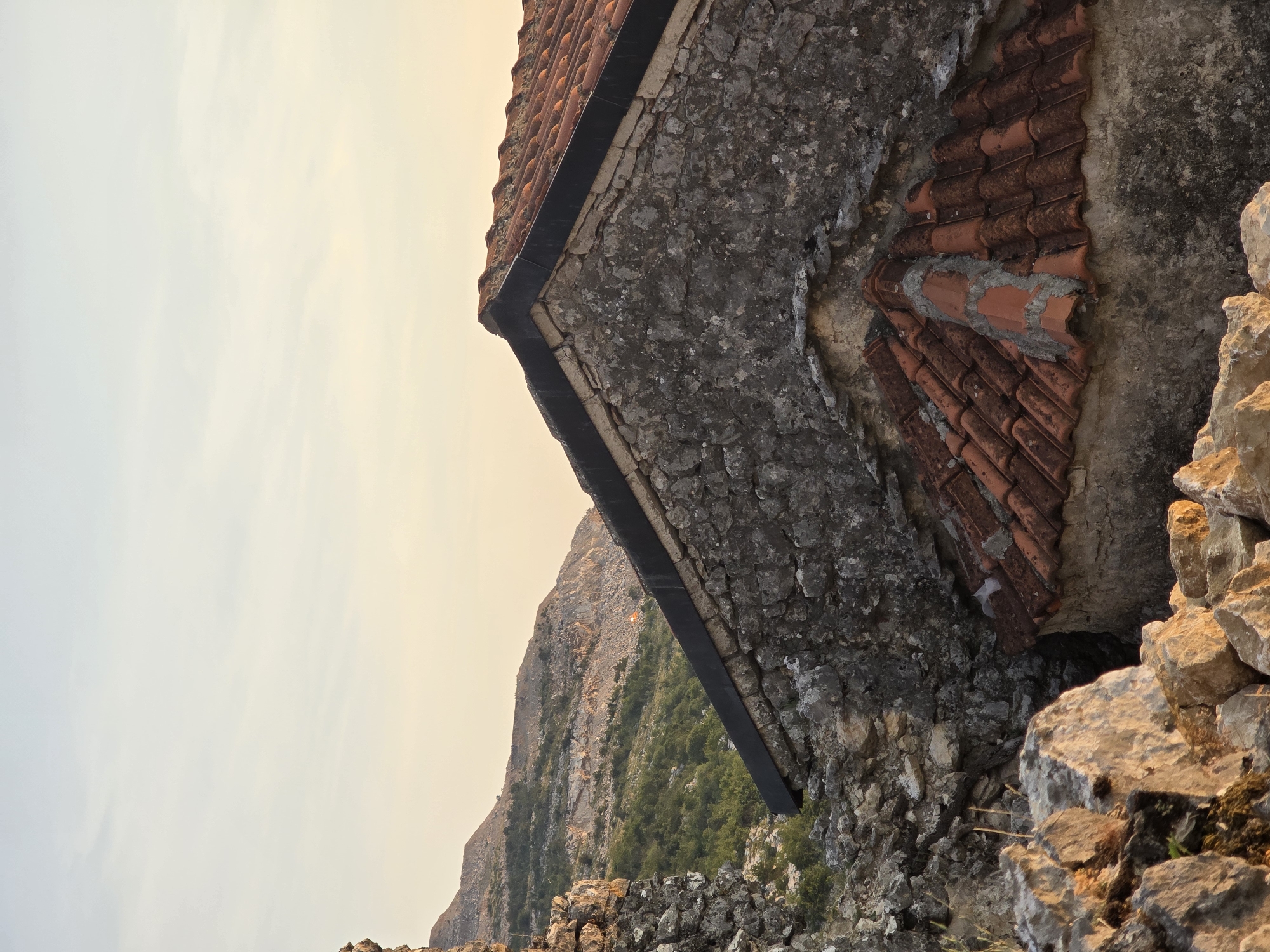 This is a close-up, eye-level, outdoor, vertical, medium shot of the roof and stone walls of a structure in Medun, Montenegro, likely part of the Medun Fortress ruins. The sky is clear and pale yellow, indicating either dawn or dusk. In the foreground, a pile of rough, light-colored stones with moss is visible. Behind them, the textured, gray stone wall of the structure is prominent, with a section of red-tiled roofing visible in the lower part of the frame. The main roof of the structure, also tiled in red, is angled upwards and occupies the center and right side of the image, with a dark trim along its edge. To the left of the structure, more stone walls are visible, with a distant view of a rocky, green mountainside under the pale sky. There are no people or animals in the image.